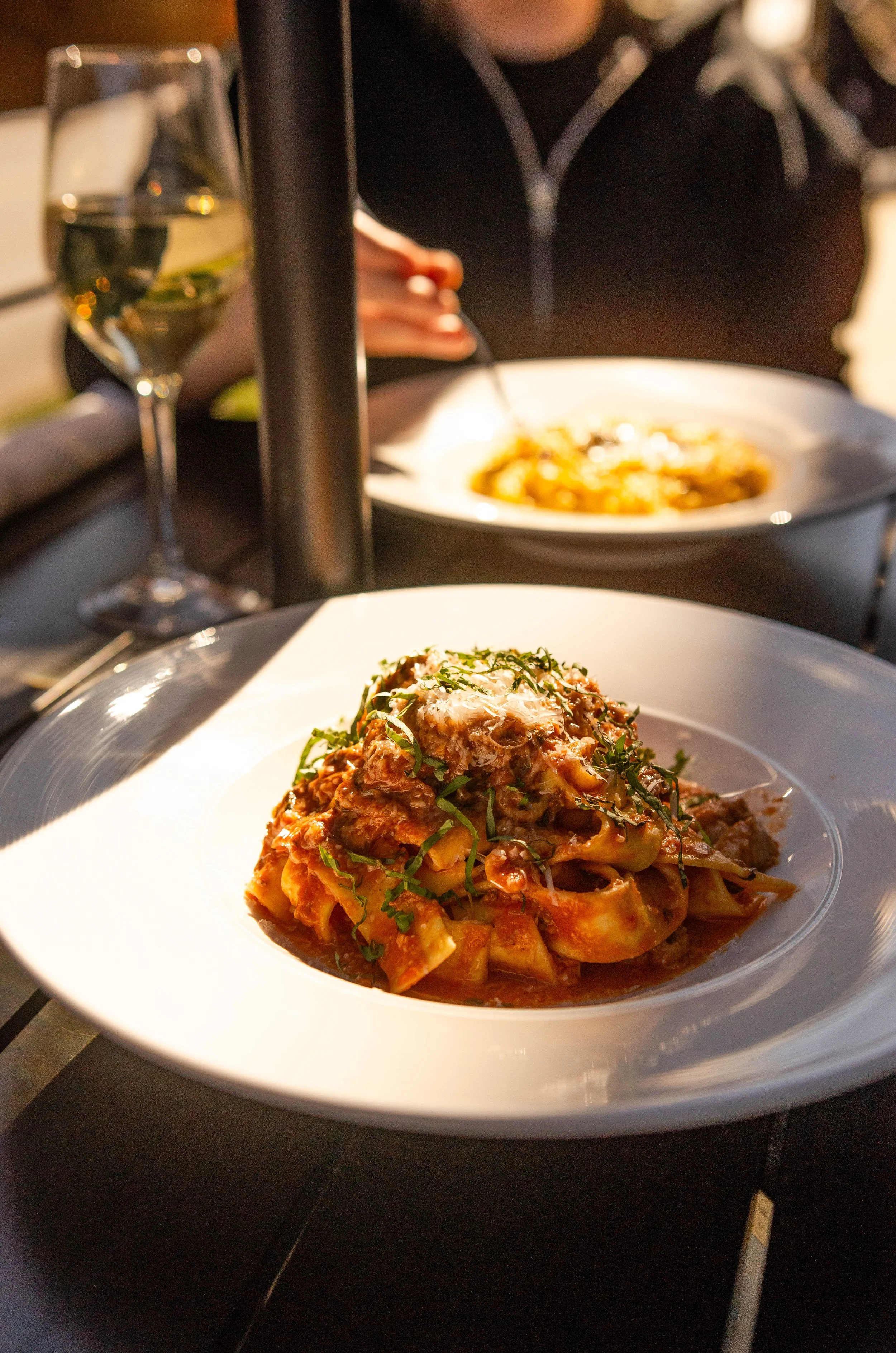A close-up of a white plate with a serving of pasta topped with meat sauce and herbs. In the background, there is a glass of white wine, and additional plates of food are partially visible along with a person in a dark shirt with stripes around the neck, dining at a restaurant.