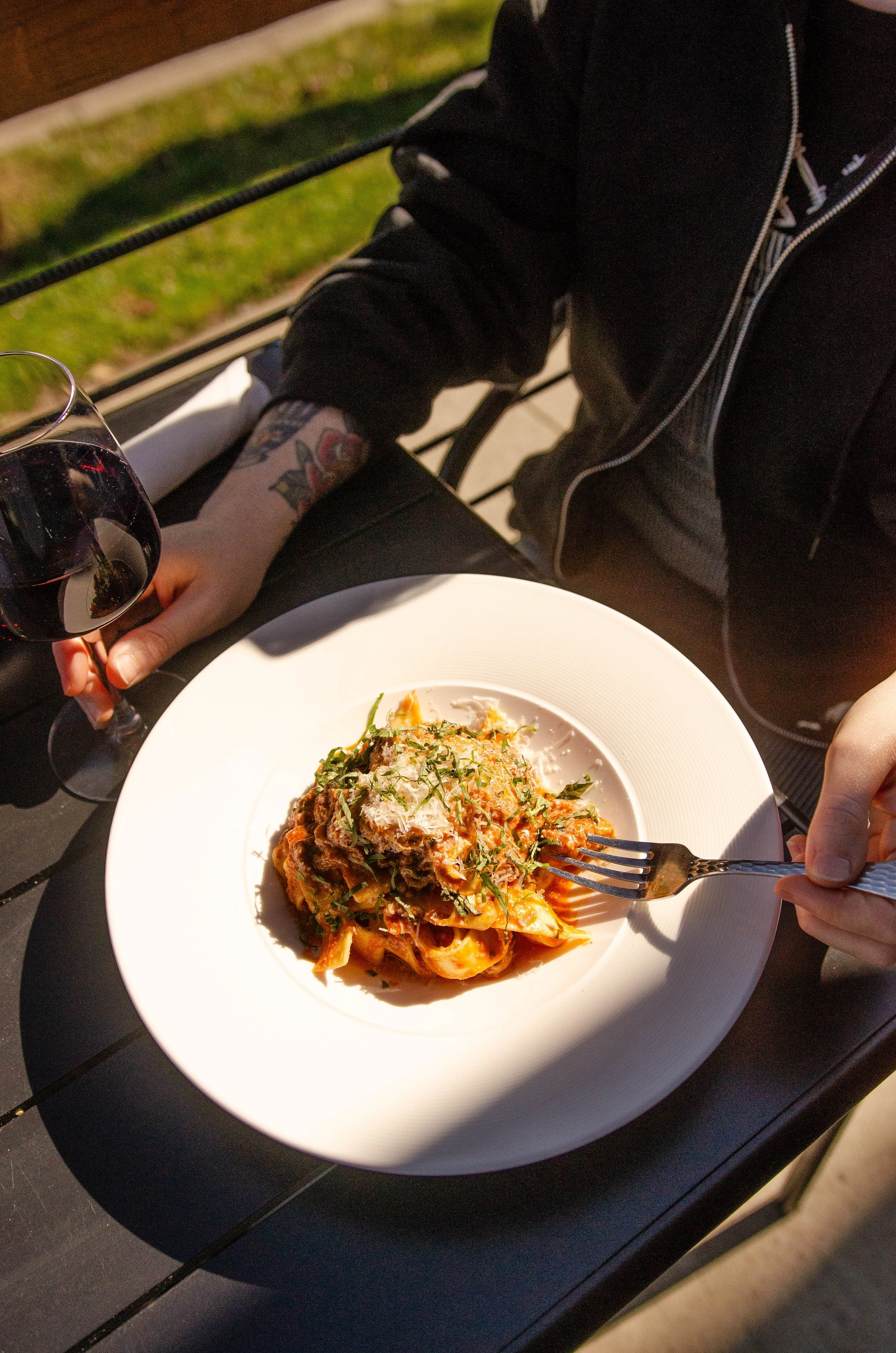A white plate of pasta topped with cheese and herbs on a black table. A person's hand is holding a fork, picking up some pasta. A glass of red wine and a person's tattooed arm are visible. The table is outdoor with grass and a wooden fence in the background.
