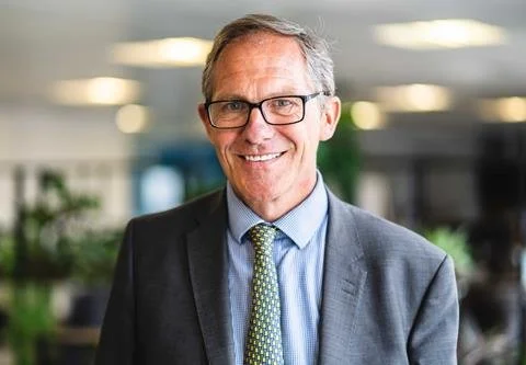 A middle-aged man in a suit and glasses smiling in an office setting.