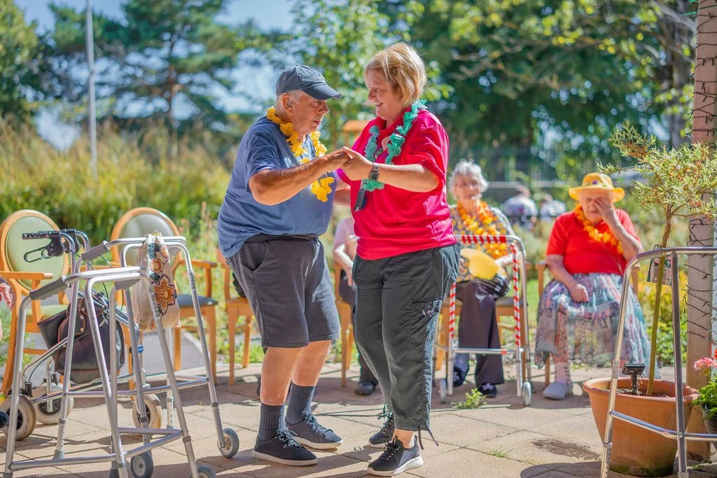 An elderly man and a young woman are dancing together outdoors surrounded by elderly women sitting in chairs, wearing leis, some with walkers, in a sunny garden.