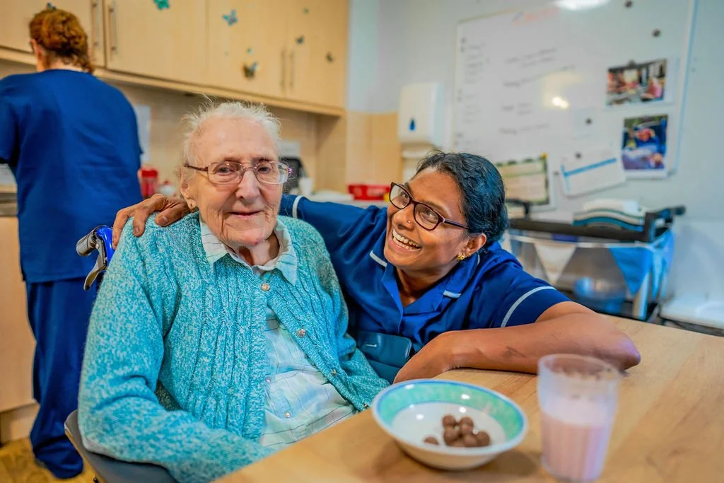 An elderly woman sitting at a table with a caregiver, both smiling; the caregiver has her arm around the woman. There is a bowl with cereal and a glass of milk or juice on the table. A person in blue scrubs is in the background of a kitchen or care f