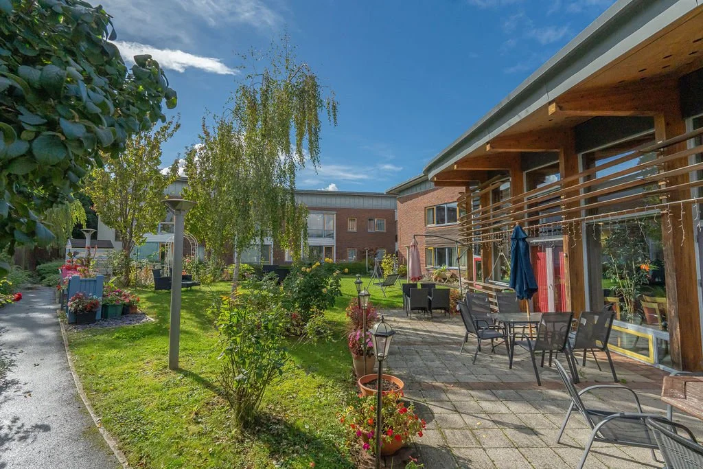 A backyard patio scene with outdoor seating, potted plants, and a grassy lawn, adjacent to a multi-story residential building with wooden balcony rails, under a partly cloudy sky.