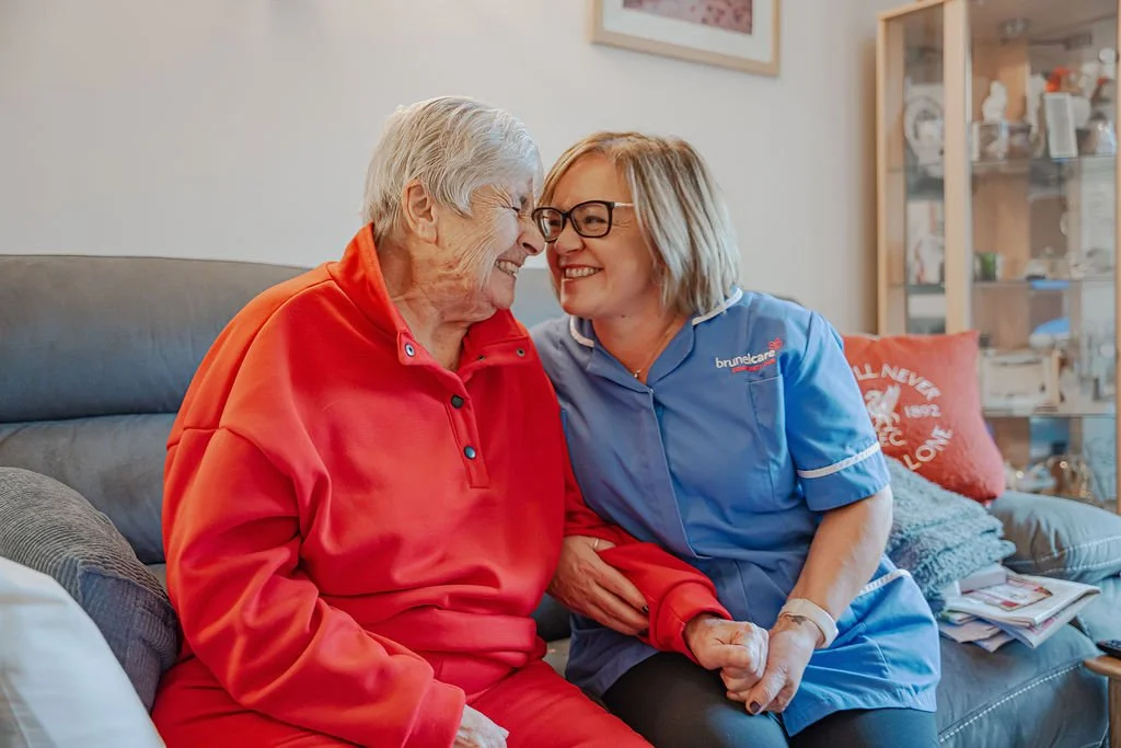 A caregiver and an elderly woman smiling and touching noses in a cozy home setting.