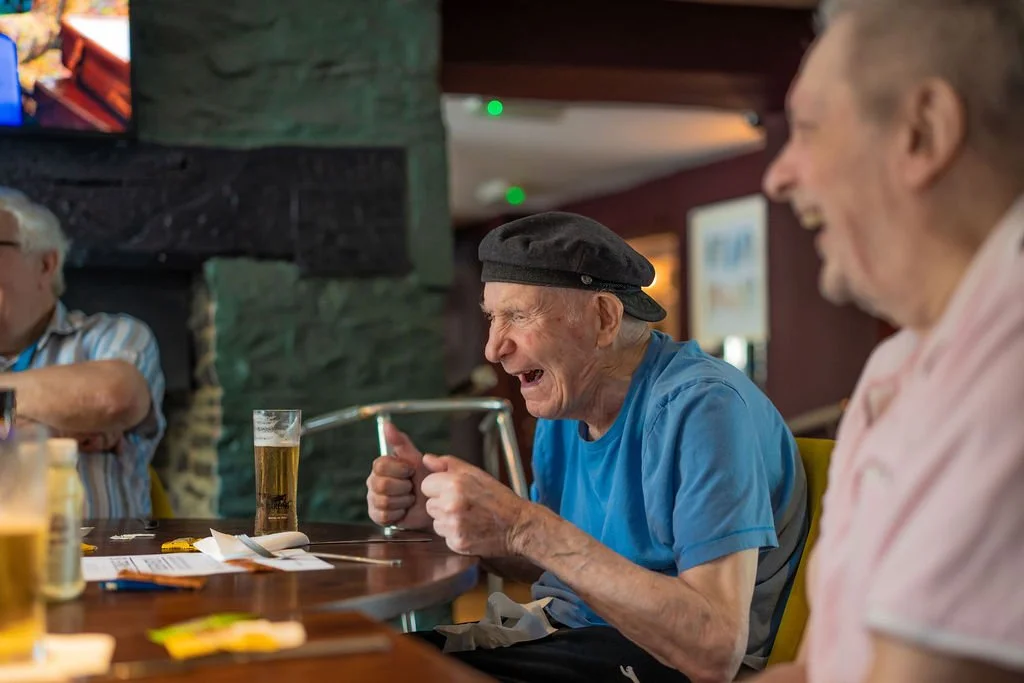 Three elderly men at a bar, laughing and enjoying drinks, with one man wearing a black beret.