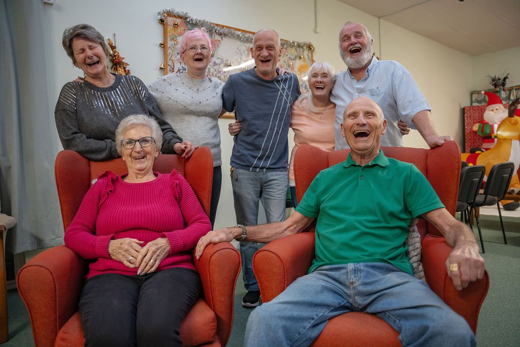 Group of elderly people celebrating and laughing, some sitting in red chairs and others standing behind them in a decorated room with Christmas ornaments and a Santa figure in the background.