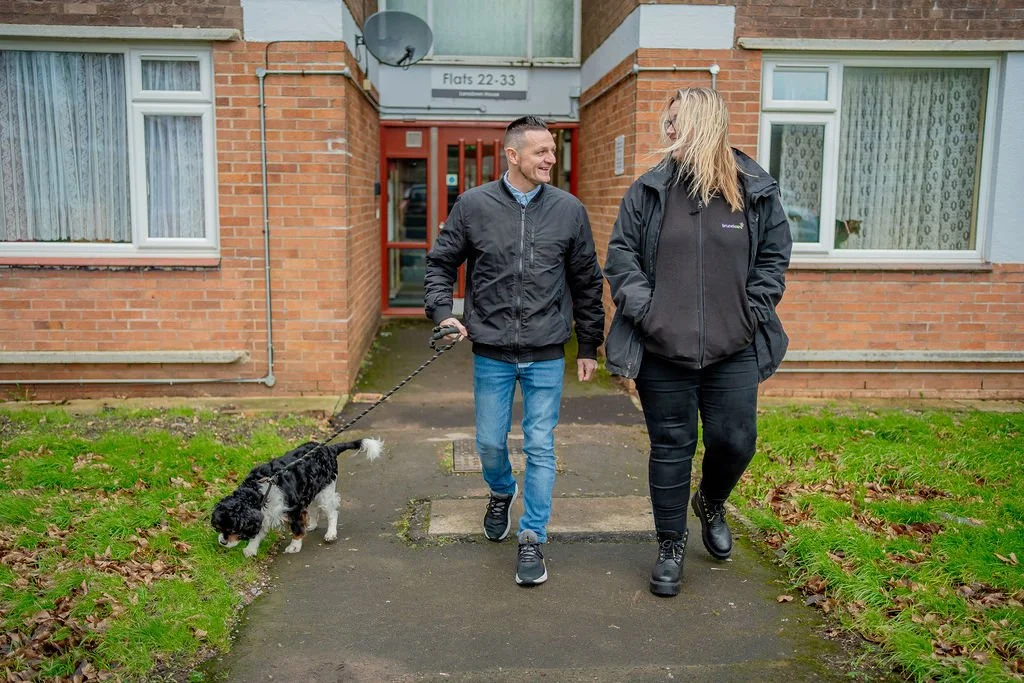 Two people walking outside an apartment building with a small dog on a leash, smiling and talking.