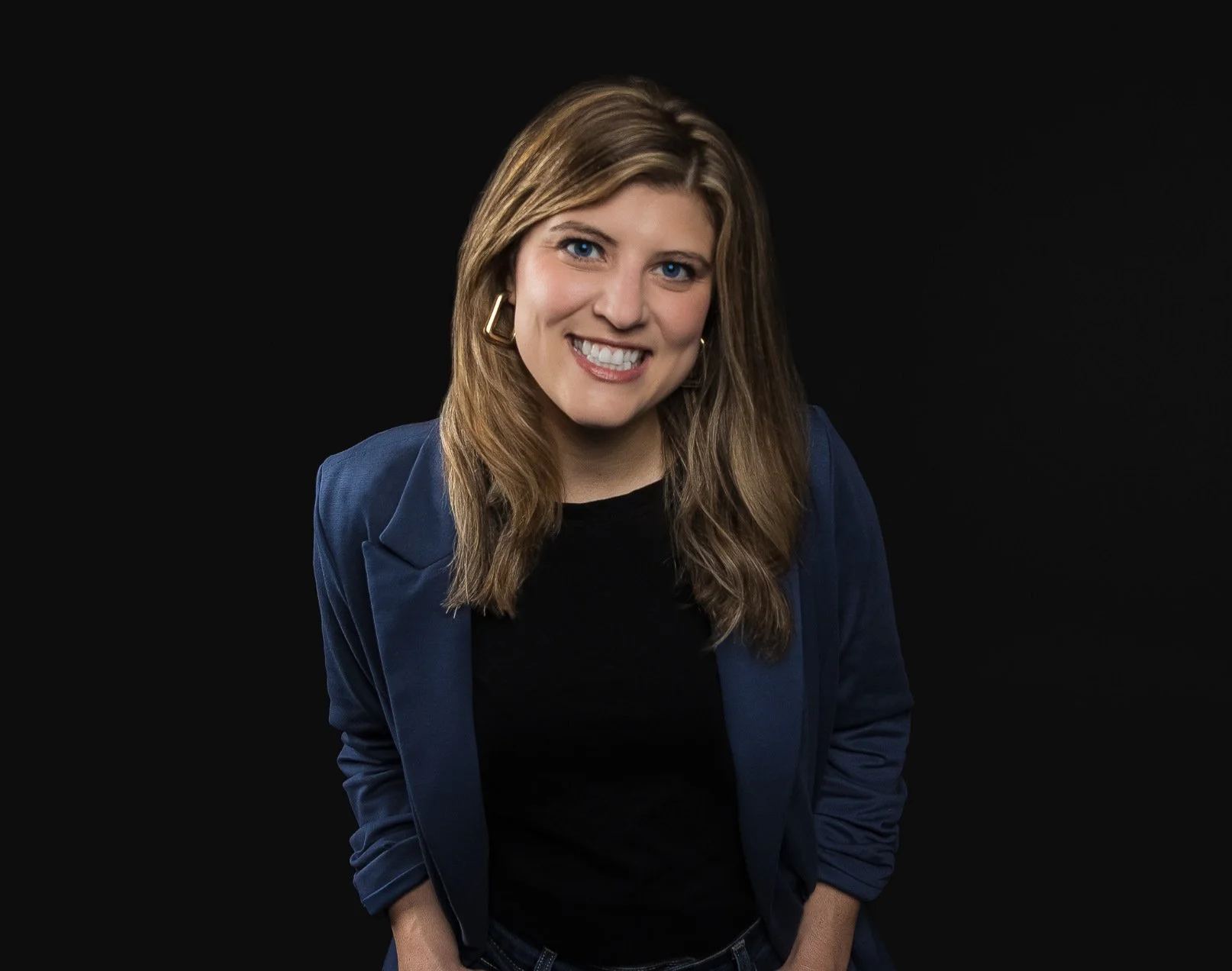 A woman with long, wavy, light brown hair, blue eyes, smiling, wearing a black top and a blue blazer, against a black background.