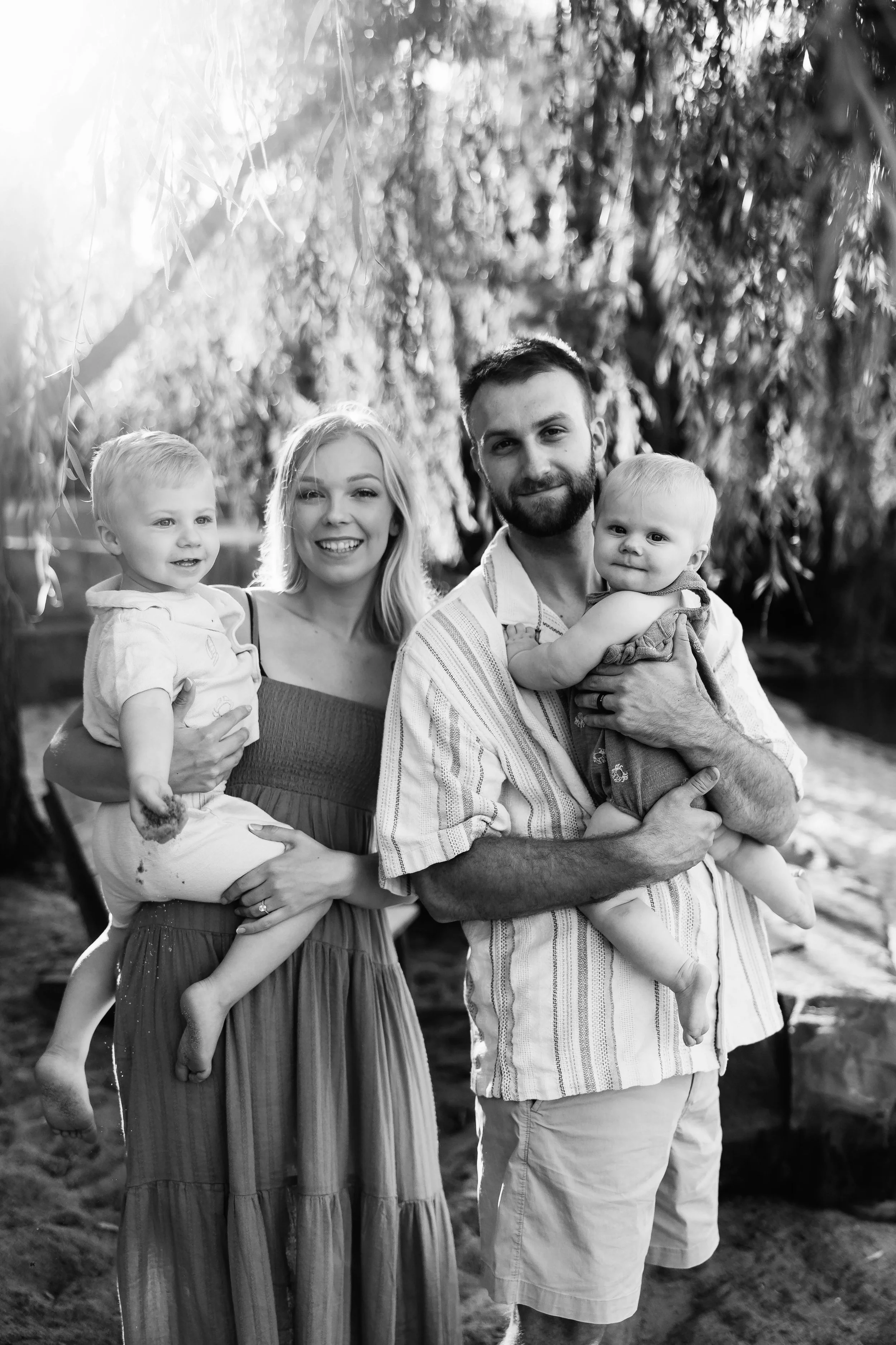 A black-and-white photo of a smiling family of four outdoors, with trees in the background. The mother is holding a young girl, the father is holding a young boy, and they are all looking at the camera.