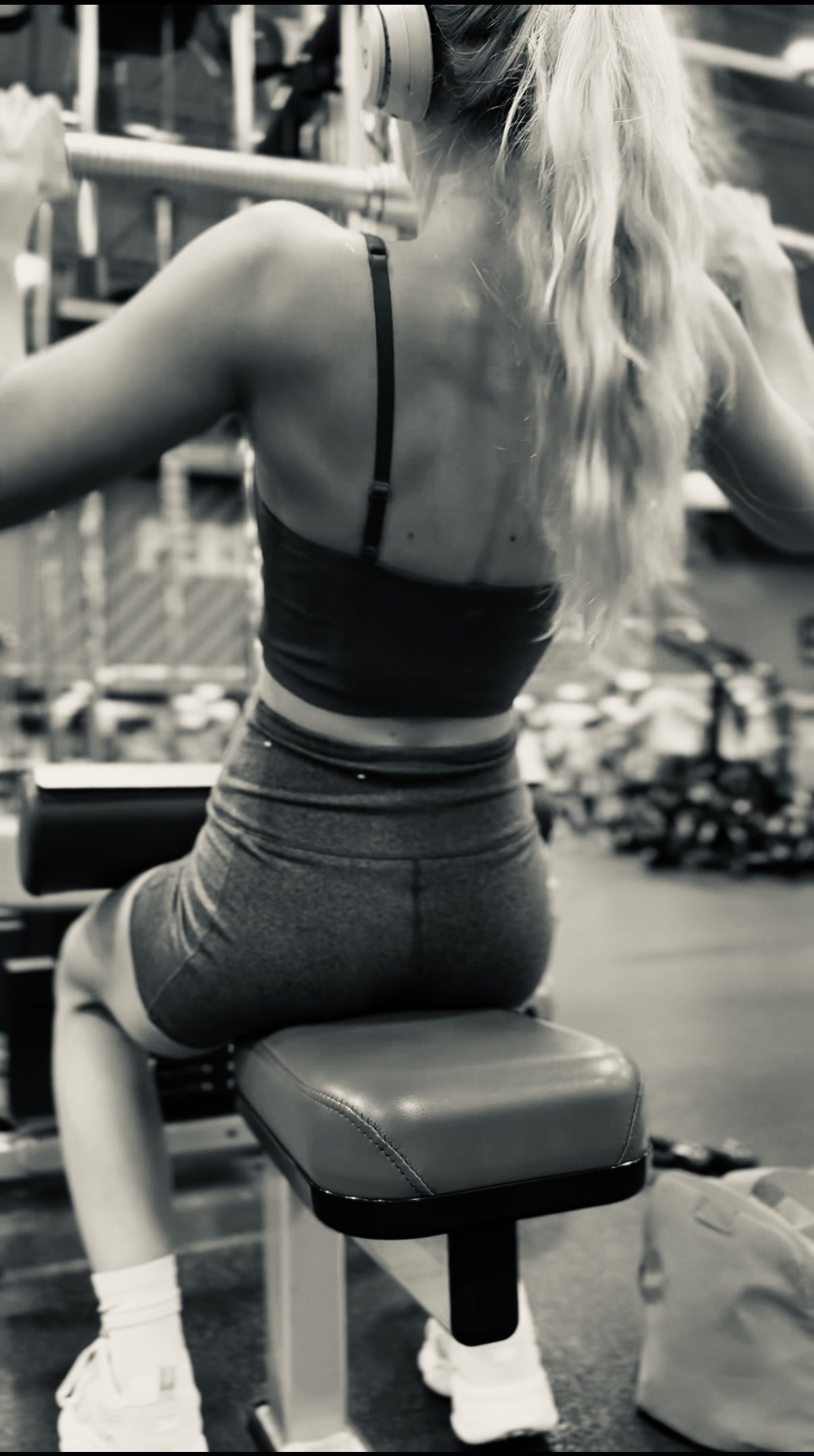A woman with long blonde hair in a ponytail sitting on a gym machine, seen from behind, in black and white.