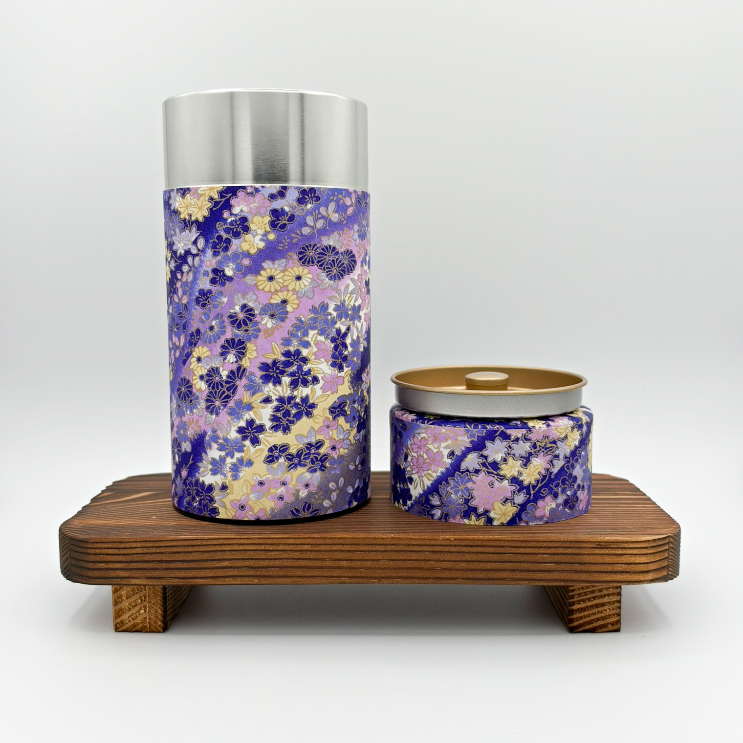 Two decorative metal containers with floral patterns on a wooden platform. The taller container is silver with a purple, yellow, and pink floral design and a silver lid. The smaller container has a similar floral pattern with a gold lid, partly open.