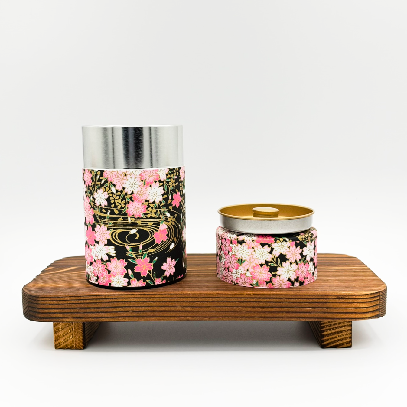 A wooden platform displaying two decorative containers with cherry blossom patterns, one tall and cylindrical with a metallic lid and one short and round with a two-tone lid, against a plain white background.