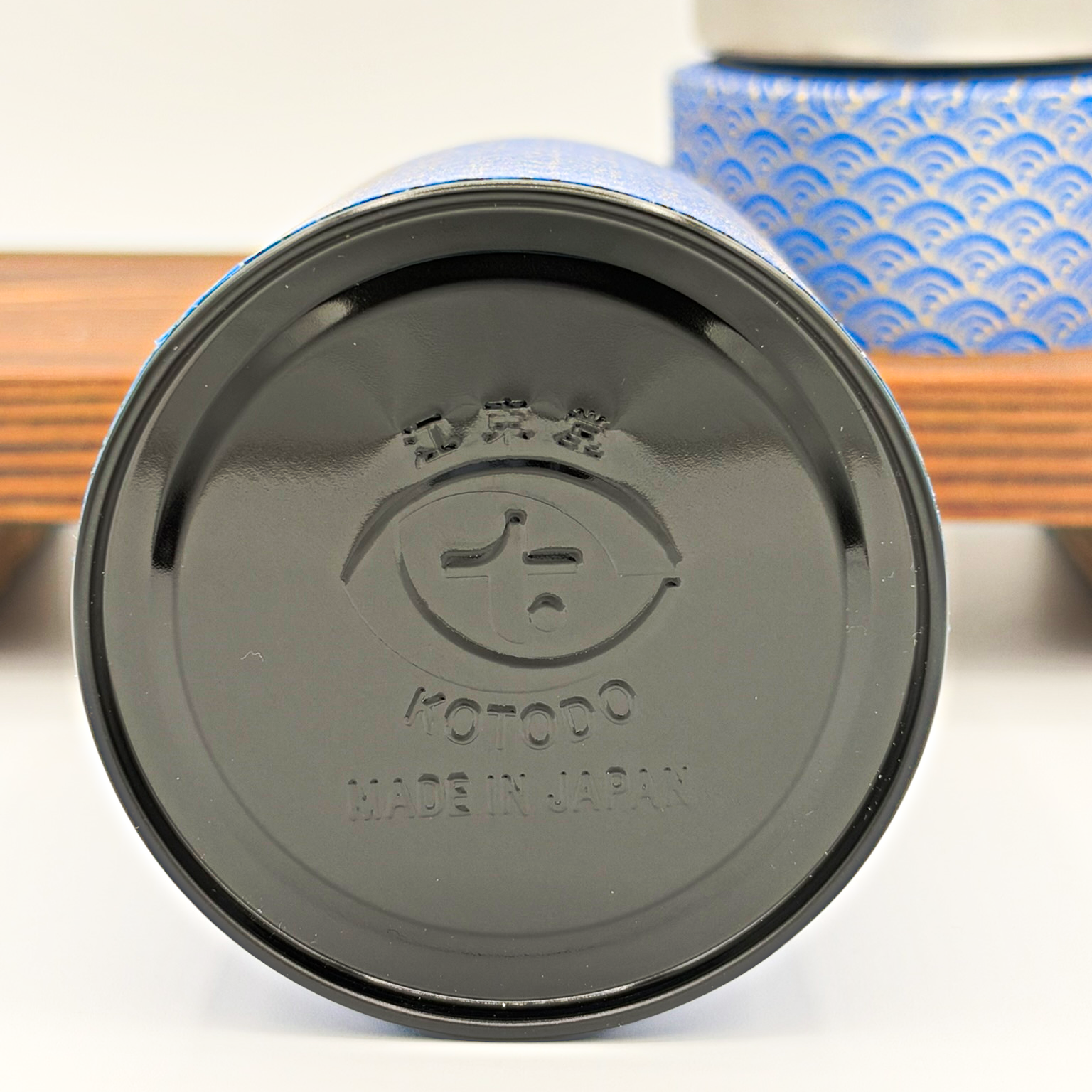 The bottom of a can with the brand name 'KOTODO' and 'MADE IN JAPAN' embossed, facing upwards. The can is placed on a white surface with a blurred background that includes a blue patterned box and a wooden surface.