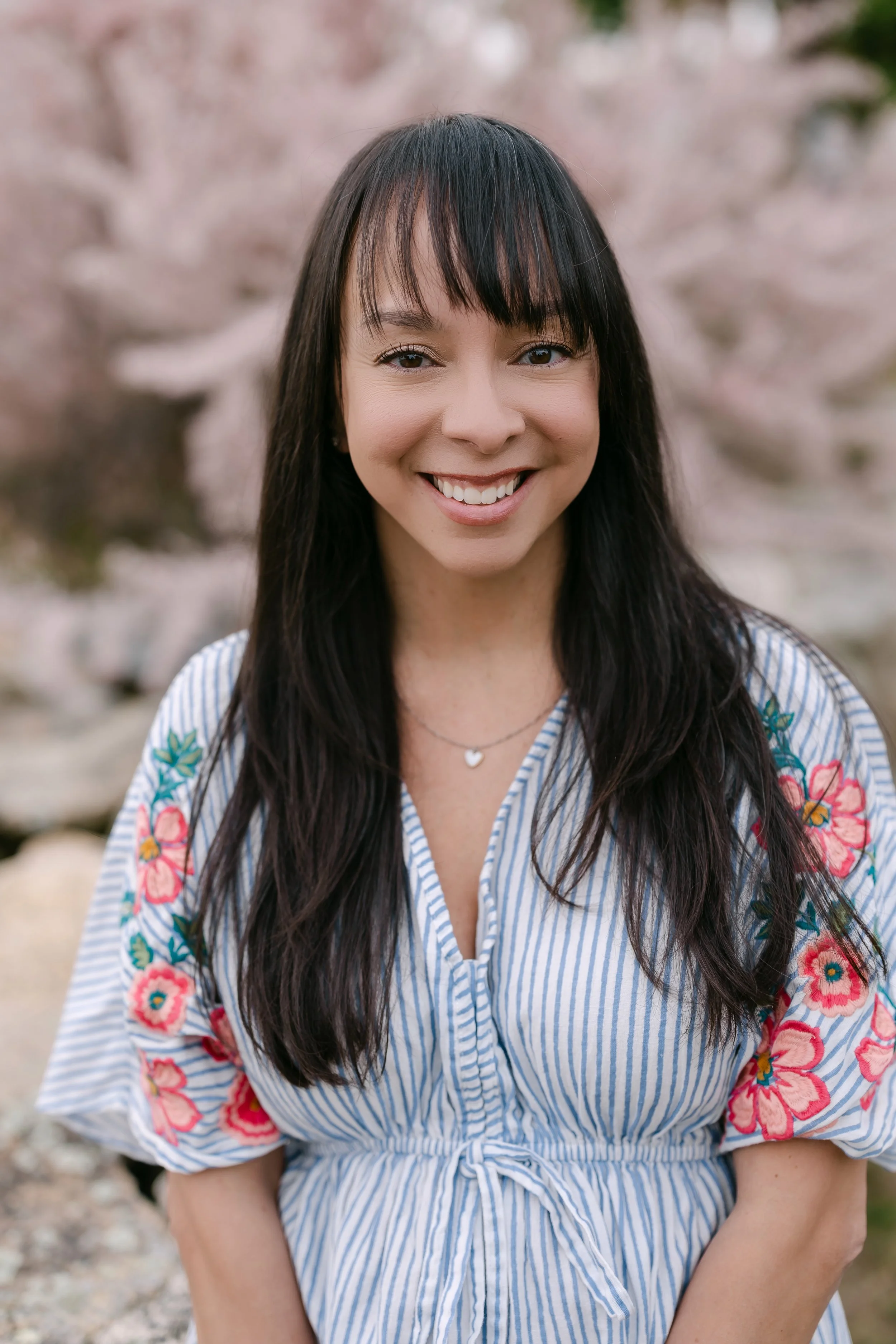 A woman with long black hair and bangs smiling outdoors, wearing a blue and white striped dress with floral embroidery on the sleeves, against a background of pink cherry blossoms.