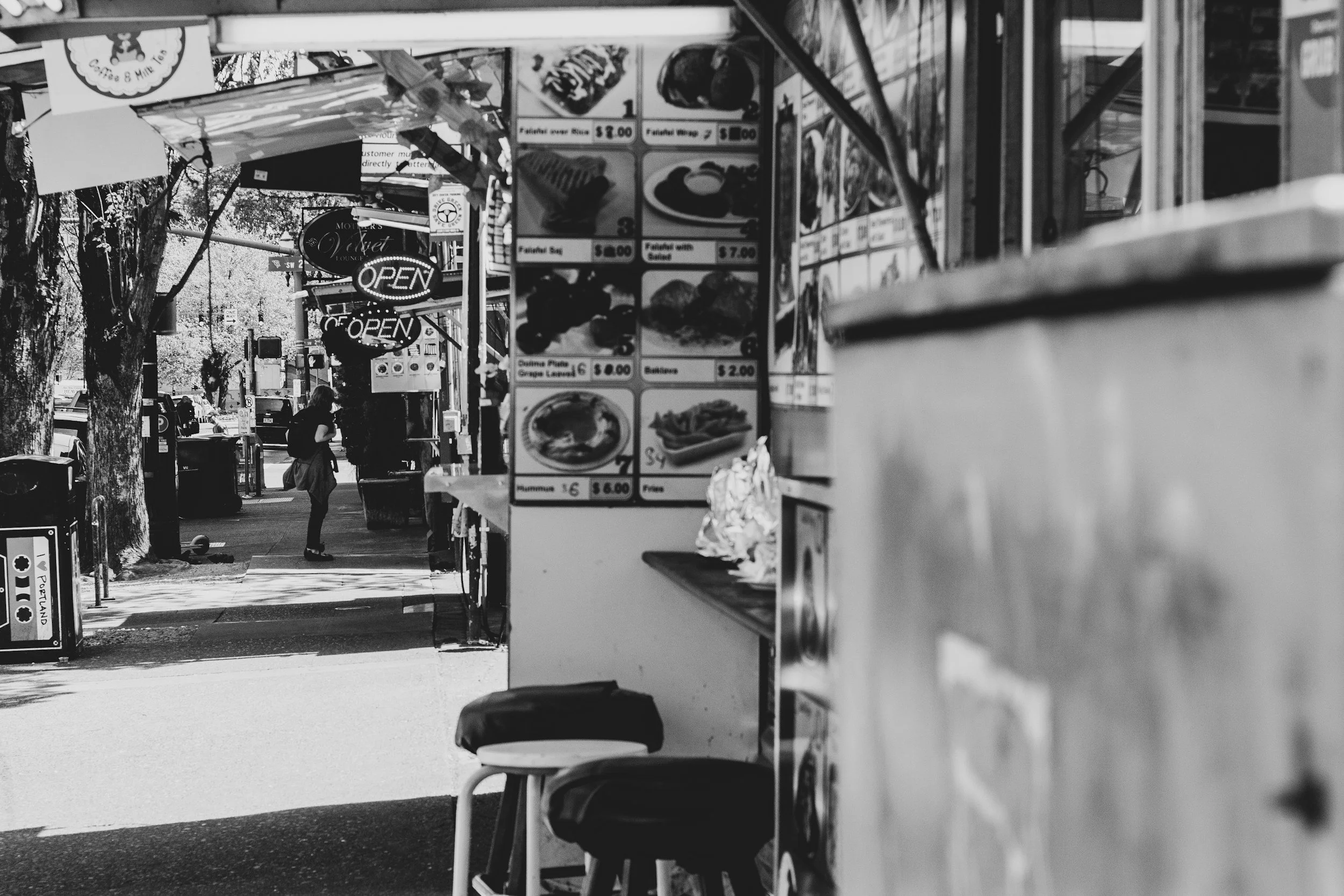 Black and white photo of a city street scene with a food stand, pedestrian, and trees in the background. The food stand has menu posters and an illuminated "Open" sign.
