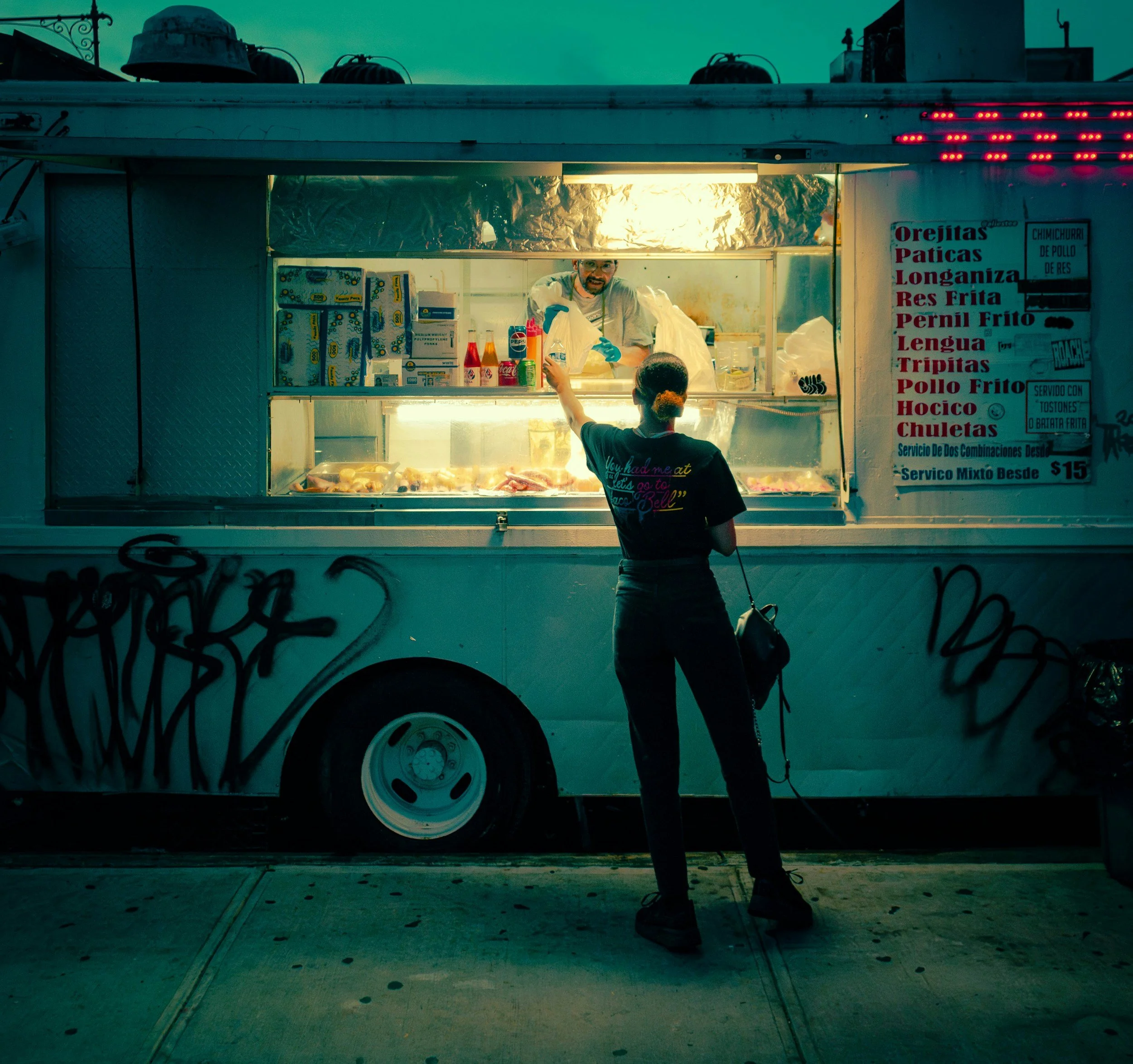 A food stand with a person serving a customer, selling fried chicken and other foods, outdoors during dusk or evening, with graffiti on the wall and pipes on the roof.