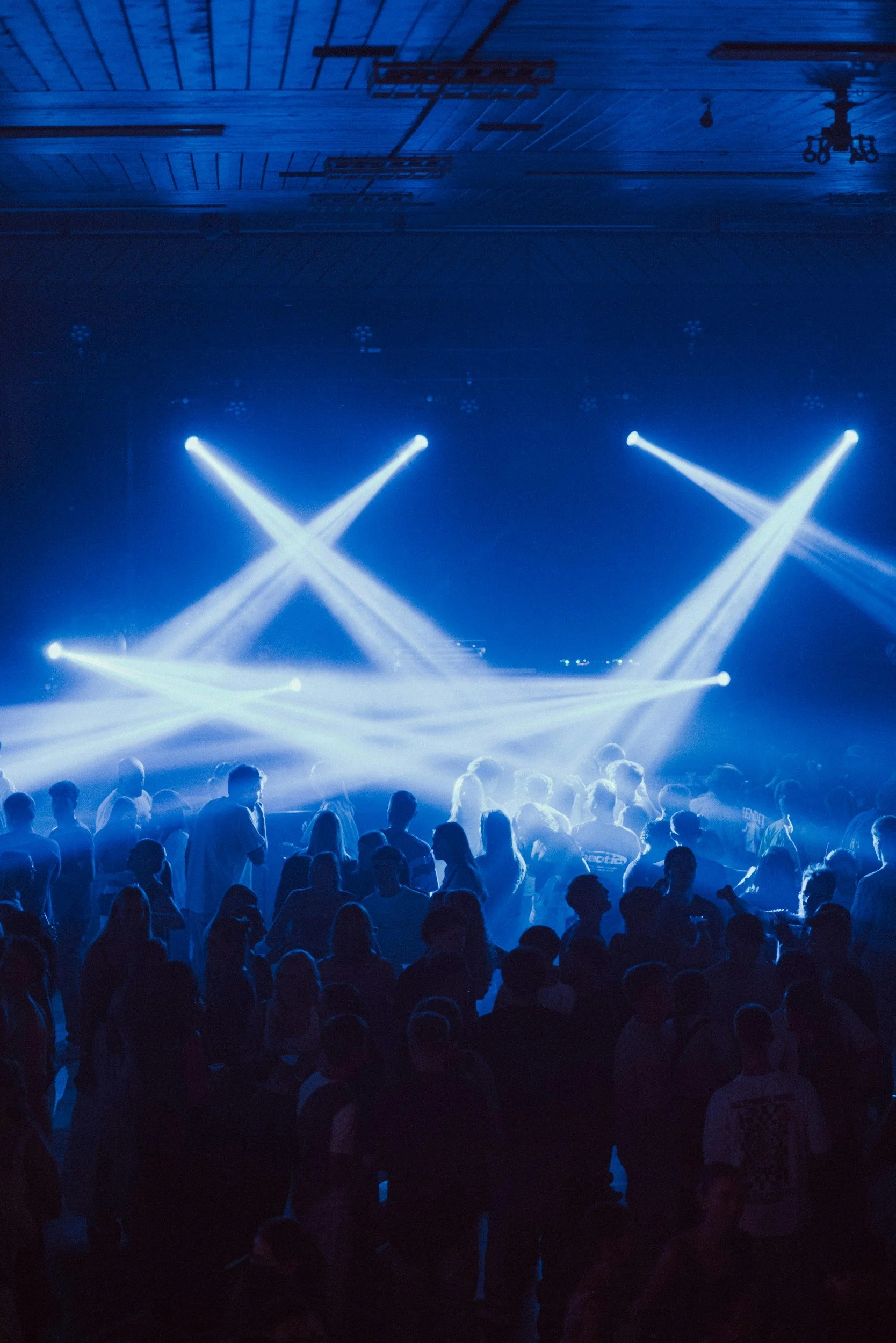 Crowd of people at concert with blue stage lights and spotlights in a large indoor venue.