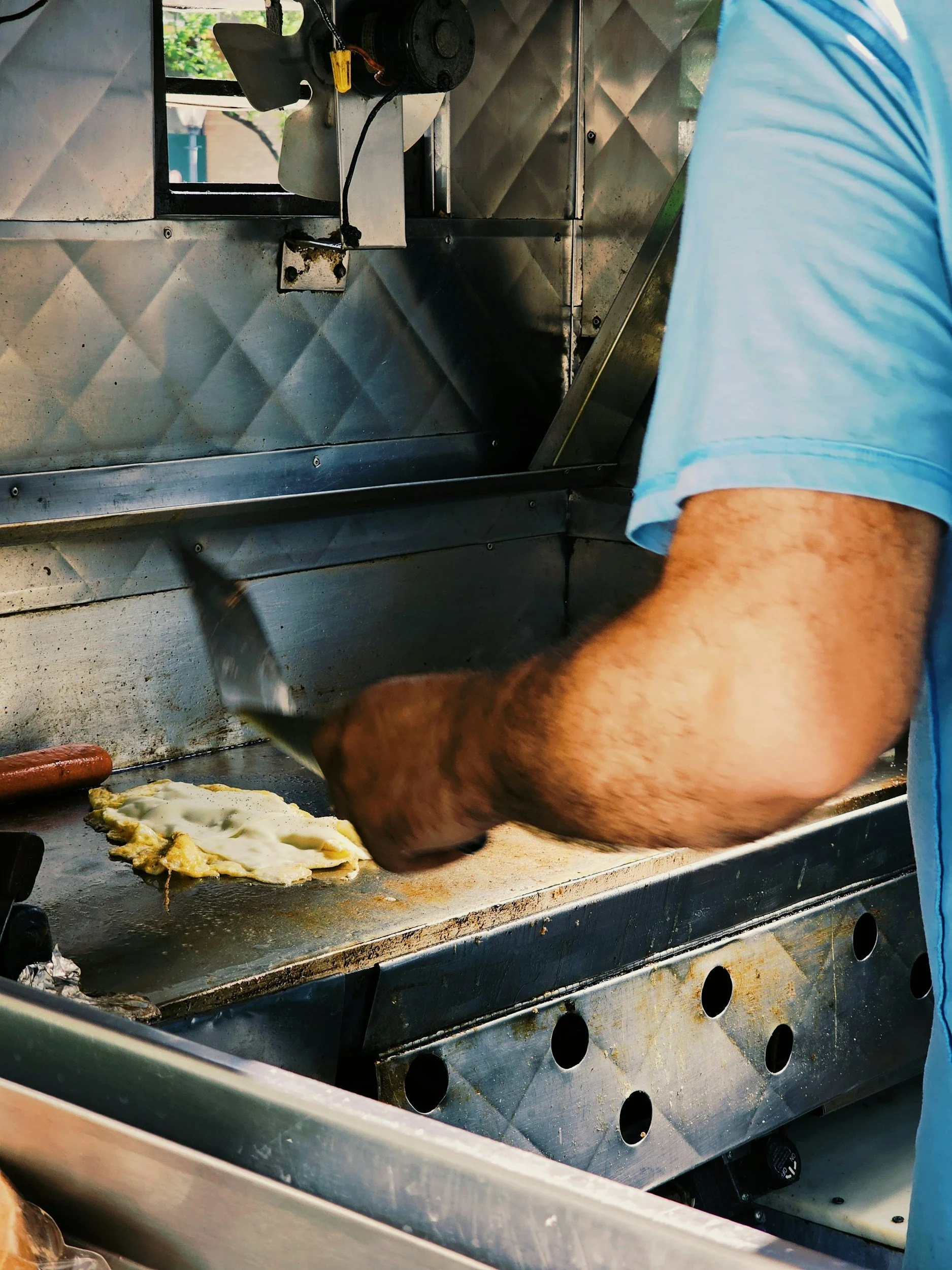 A person wearing a light blue shirt cooking eggs on a flat-top grill, with a spatula in hand. The grill has a stainless steel surface, and some eggs are present on it.