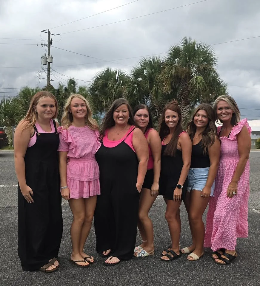 Seven women standing side by side outside, with palm trees and cloudy sky in the background, smiling at the camera.