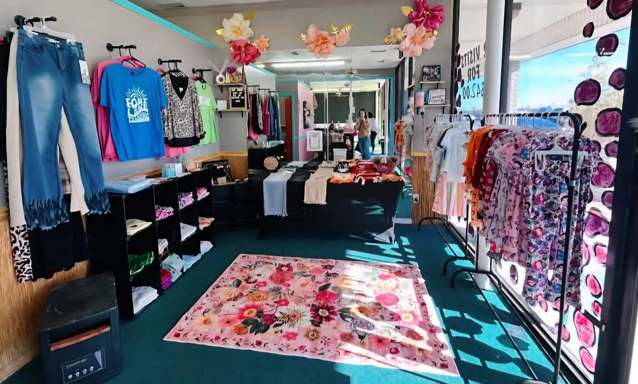 Inside a small clothing boutique with racks of colorful clothing, tables with folded items, and pink floral decor on the ceiling. A woman is visible in the background near the entrance.