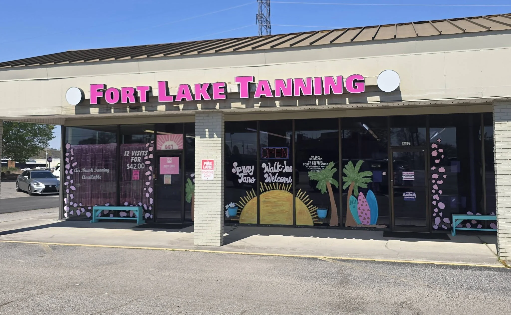 storefront with a sign that reads 'Fort Lake Tanning' in pink letters, decorated with painted palm trees and surfboards, and featuring an 'Open' sign in the window.