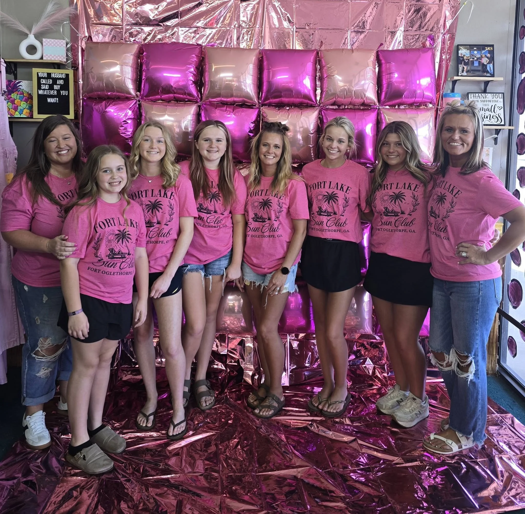 Group of eight women standing in front of a pink and purple metallic backdrop, all wearing matching pink T-shirts that say 'Fort Lake Sun Club Fort Oglethorpe, GA,' smiling and posing for the photo. The women are of various ages, some with long hair, some with curly hair, and are dressed casually.