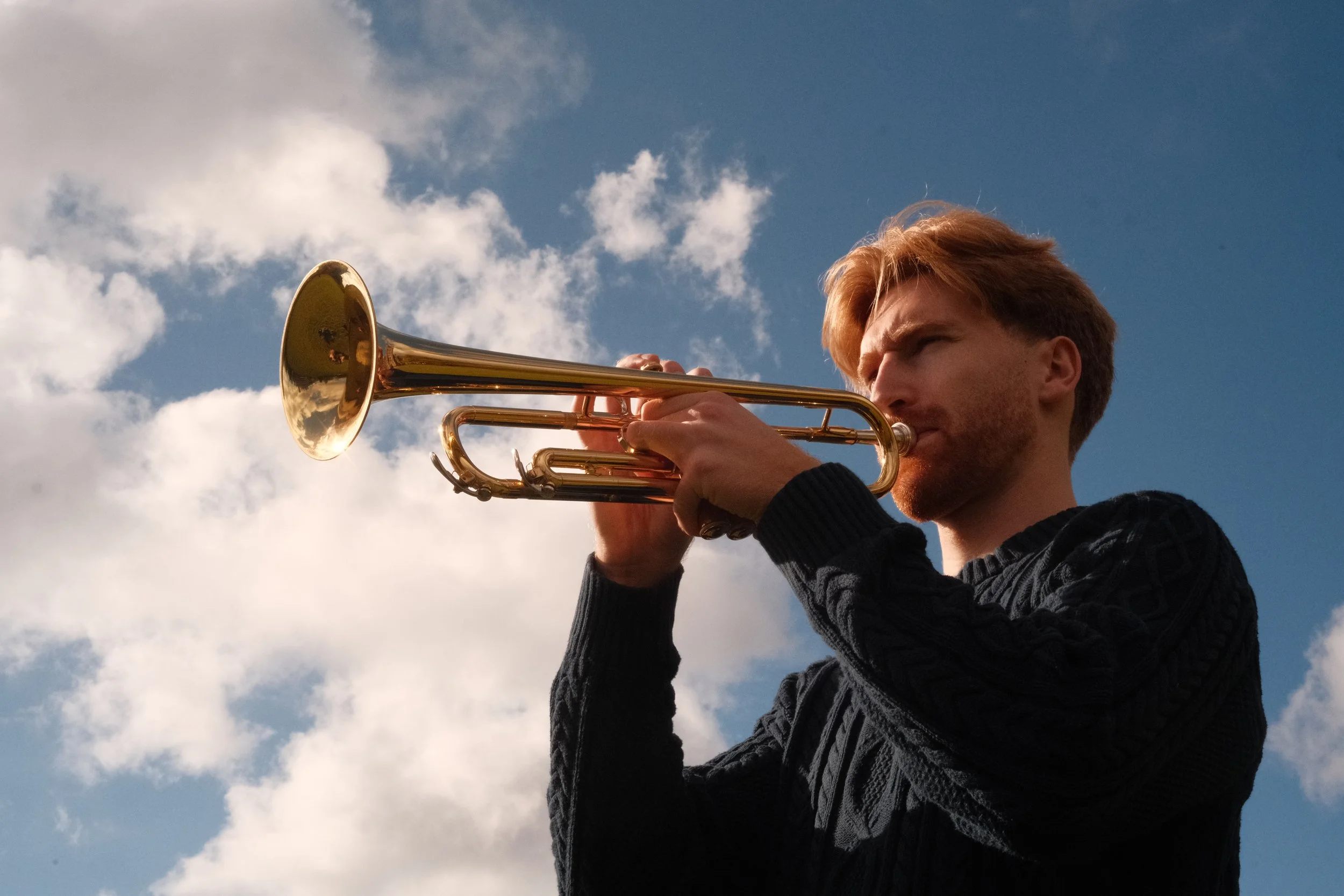 A young man with red hair playing a trumpet outdoors against a partly cloudy sky.