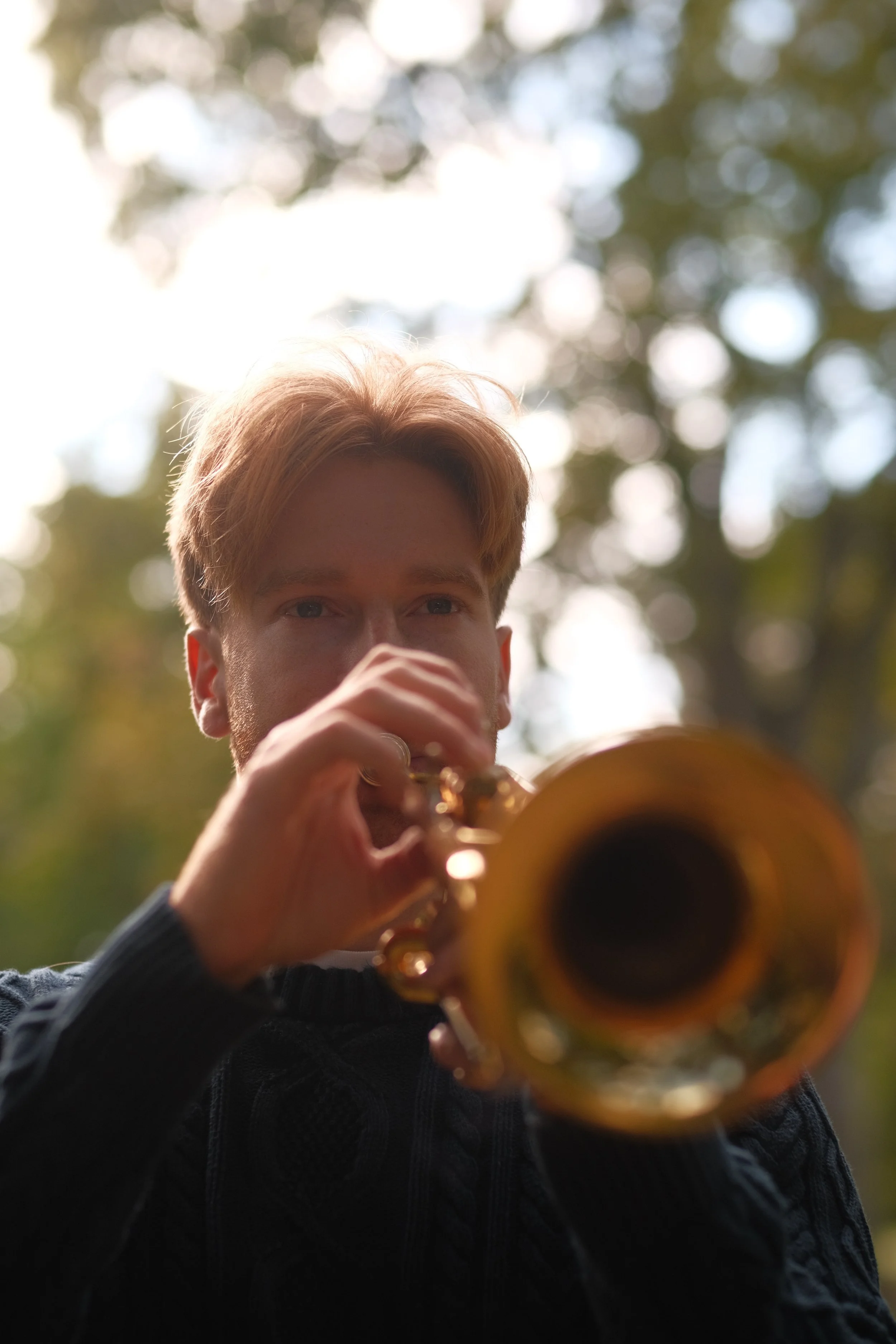 A young man with light brown hair playing a saxophone outdoors, with a blurred background of trees and sunlight.