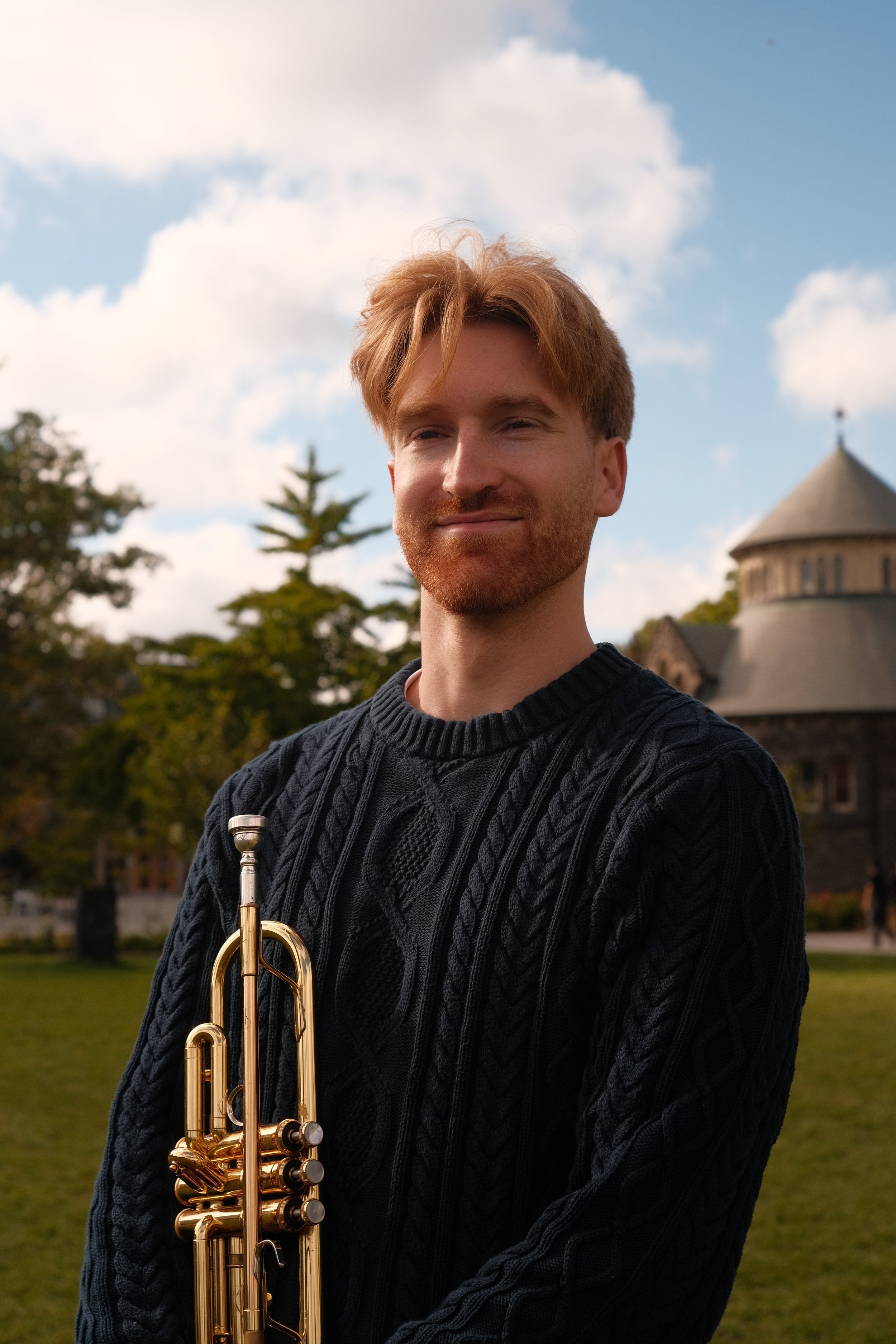 A man with red hair and a beard holding a trumpet outdoors, with trees and a stone building with a conical roof in the background.
