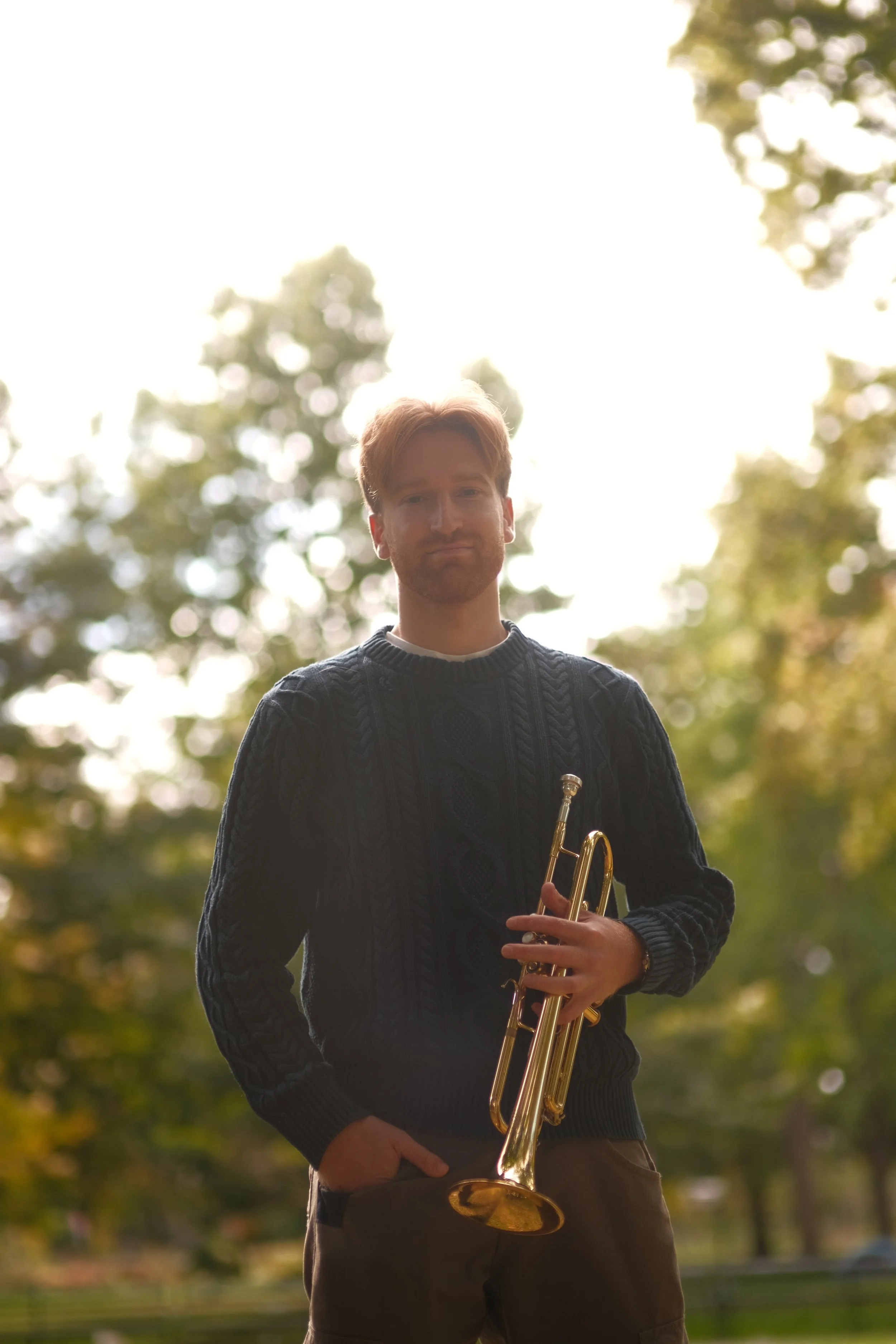 A man with red hair and a beard holding a trumpet, standing outdoors with trees and sunlight in the background.
