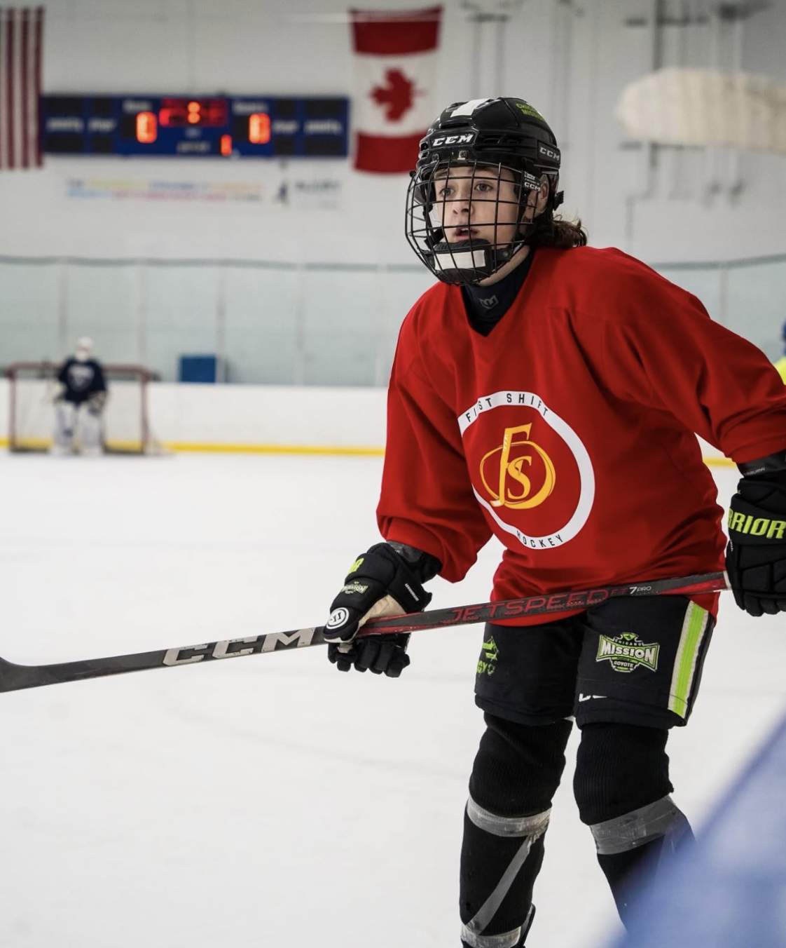 A young hockey player wearing a red jersey and black helmet with cage, holding a hockey stick, on an ice rink with a goalie in the background and a Canadian flag hanging above.