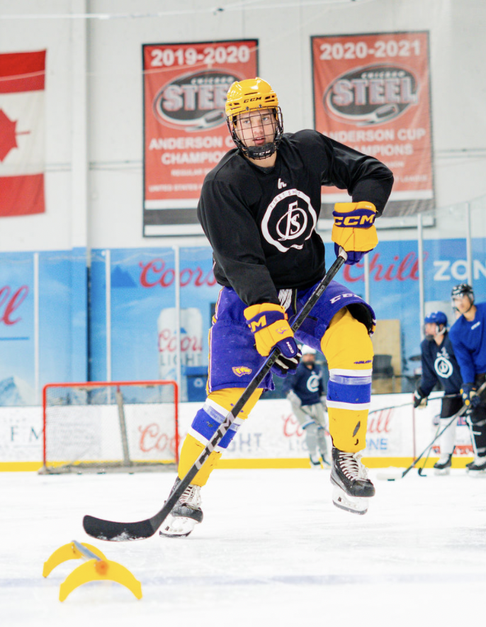 Hockey player practicing with a yellow guard on the ice rink, wearing a yellow helmet, black jersey, blue and yellow socks, yellow gloves, and holding a hockey stick.