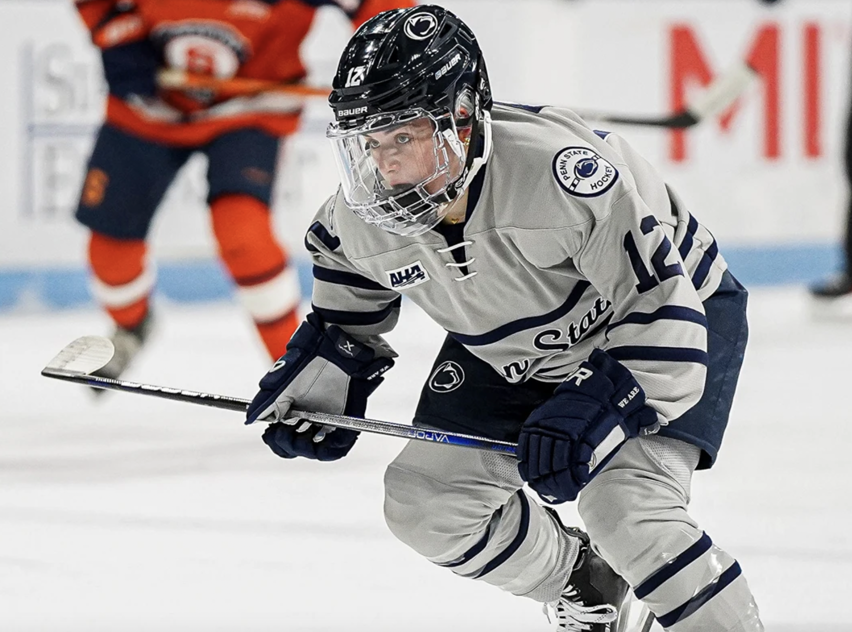 A young male ice hockey player in a gray and navy uniform with the number 12, wearing a black helmet and holding a hockey stick, on the ice rink.