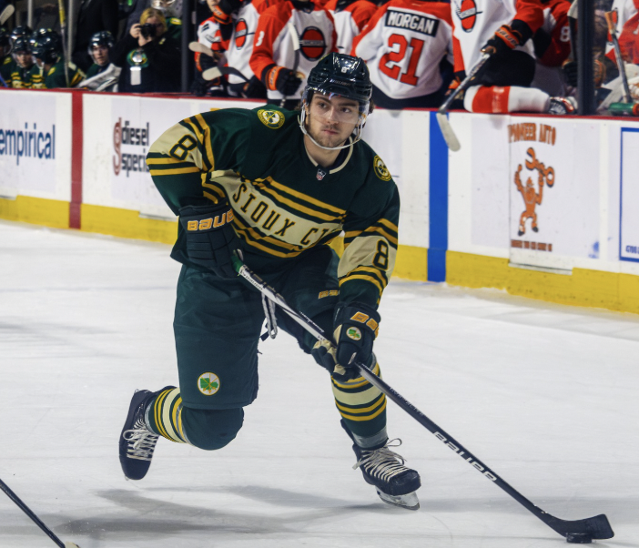 An ice hockey player in a green and yellow uniform skating on the ice during a game, with players on the bench in the background.
