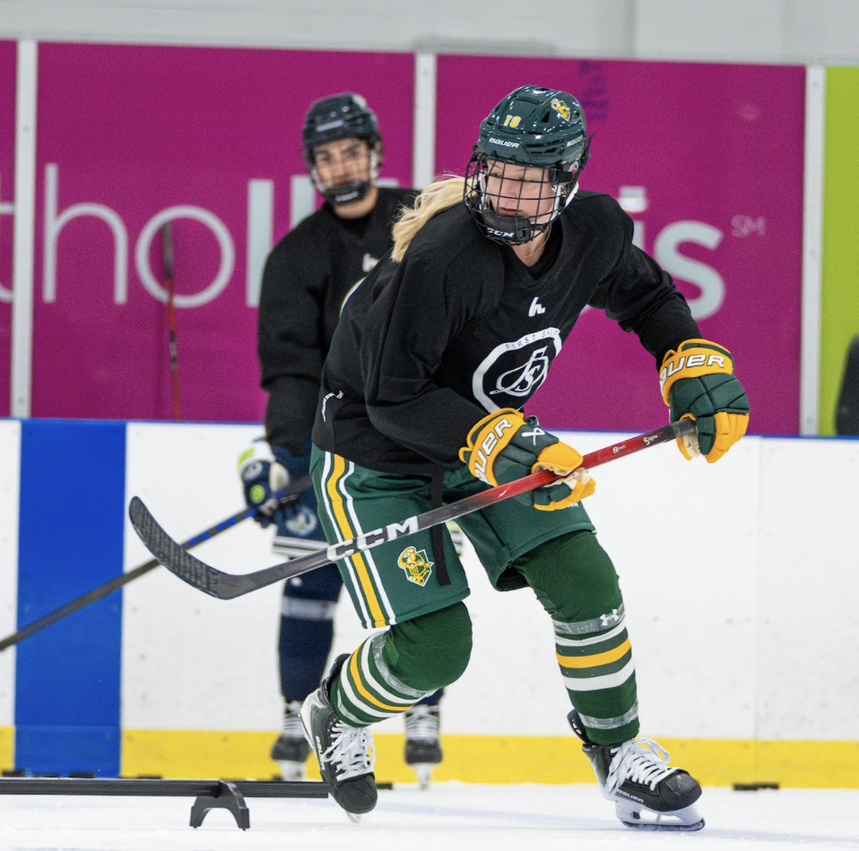 Female ice hockey player in black jersey and green uniform training on ice rink with coach in background.