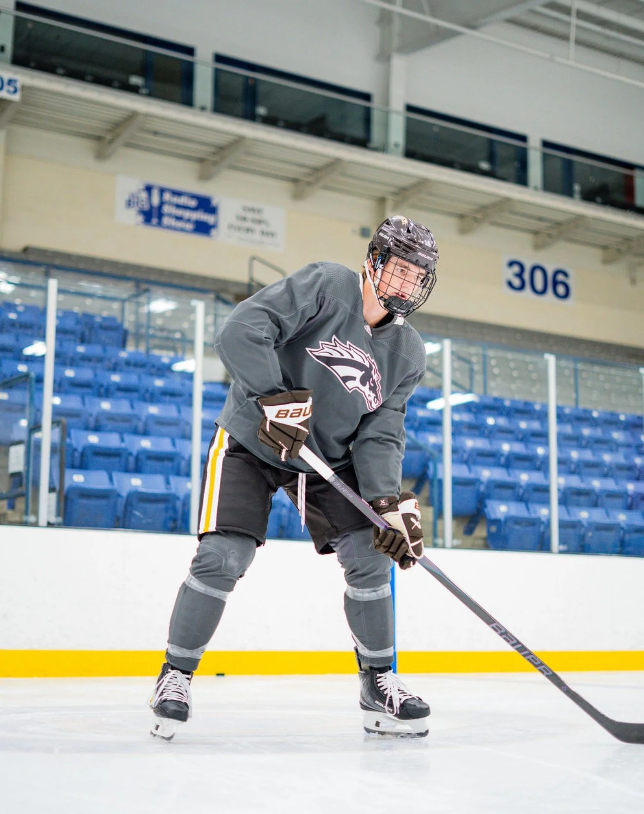 A male ice hockey player on the rink, wearing a gray jersey with a stylized animal head logo, black hockey pants with white and yellow stripes, black gloves, a black helmet with a faceguard, and holding a hockey stick, positioned in a ready stance for play.