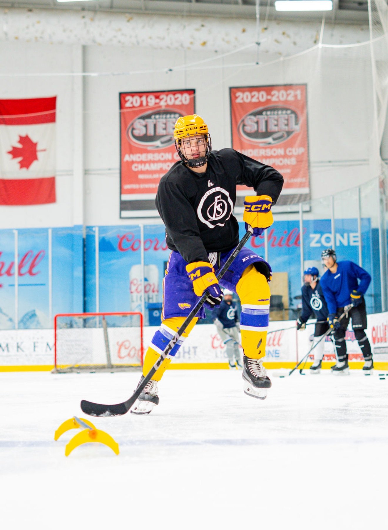 An ice hockey player wearing a yellow helmet and black jersey practicing stickhandling on the ice, with a training aid attached to their stick.