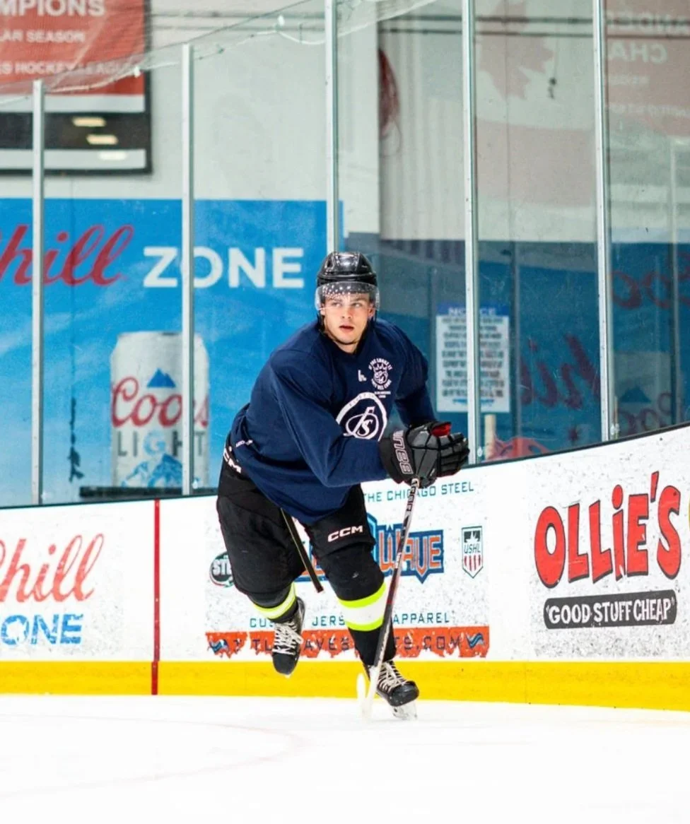 A young male ice hockey player in a dark blue jersey, black shorts with yellow stripes, and black helmet, skating on the ice in an indoor rink, holding a hockey stick and appearing focused.