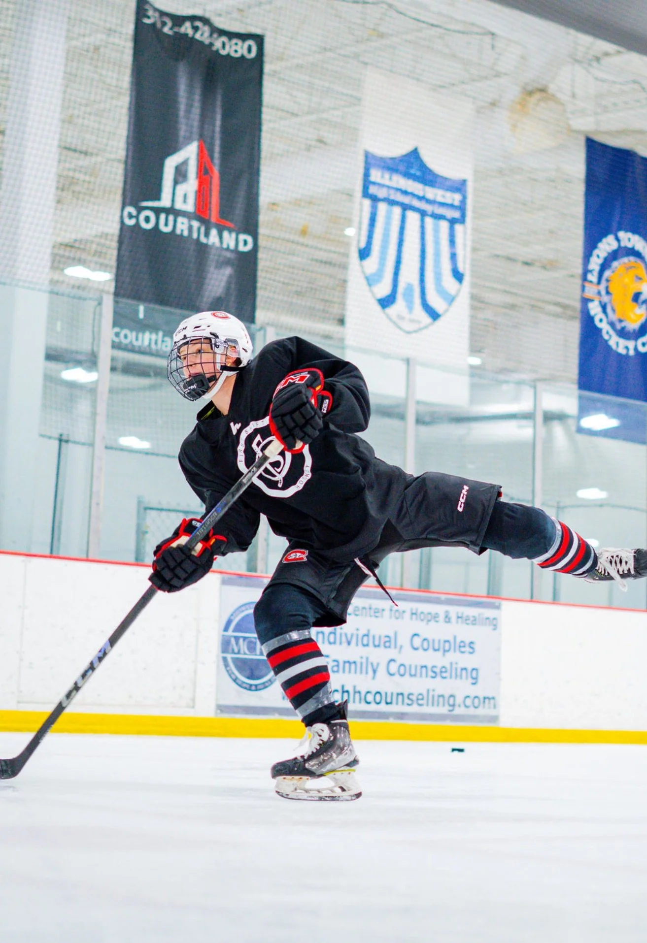 An ice hockey player in black gear and a white helmet skating on the ice rink, holding a hockey stick, with banners hanging from the ceiling in the background.