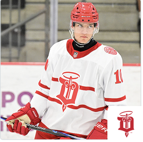 Young male ice hockey player in a white and red jersey with a red helmet holding a hockey stick, on an ice rink.