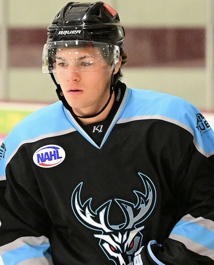 Young male ice hockey player wearing a black and blue jersey with a deer logo, helmet, and visor, on an ice rink.