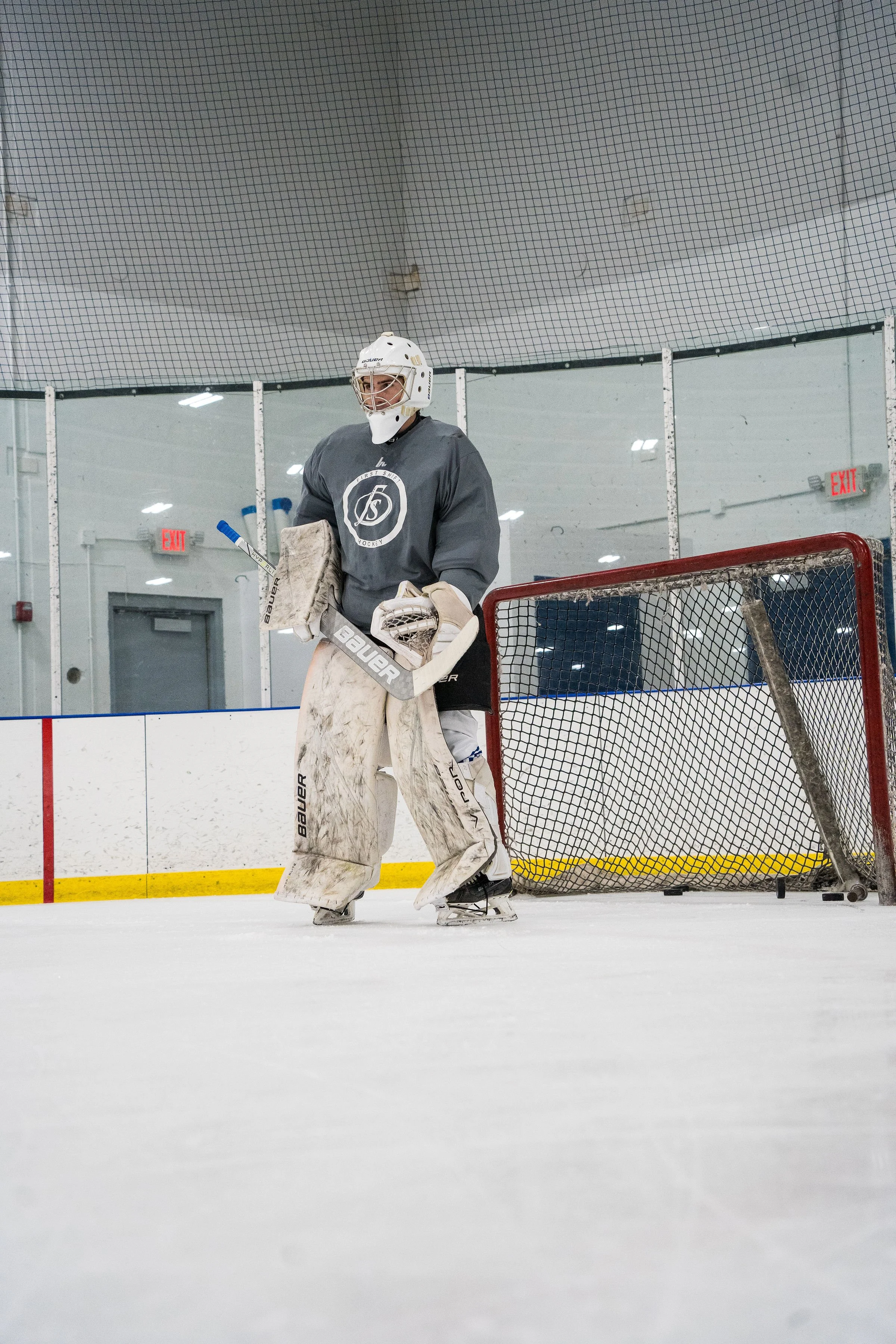 An ice hockey player standing on the rink, wearing a grey jersey, white goalie pads, and a white helmet with a face cage, holding a goalie stick and standing near the goal net, with an empty indoor ice rink in the background.
