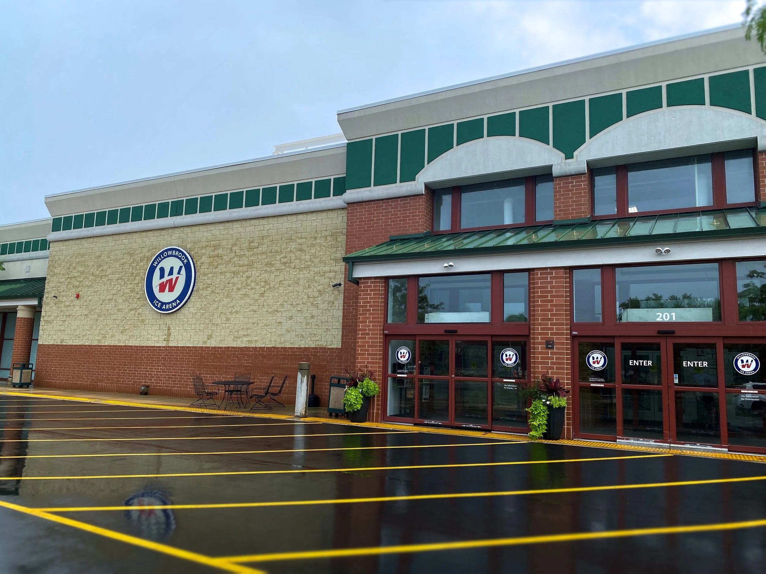 The exterior of a Willowbrook Ice Arena building with brick and stucco walls, green accents, and large windows, with a parking lot in the foreground.