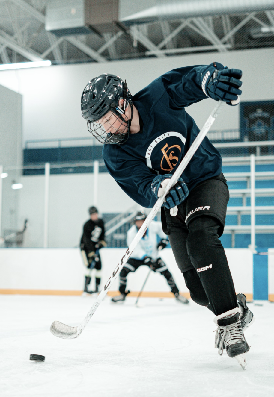 A young man in hockey gear, including a helmet, gloves, and jersey, bends down to hit a puck with his hockey stick during a practice or game on an indoor ice rink. Two other players are visible in the background.