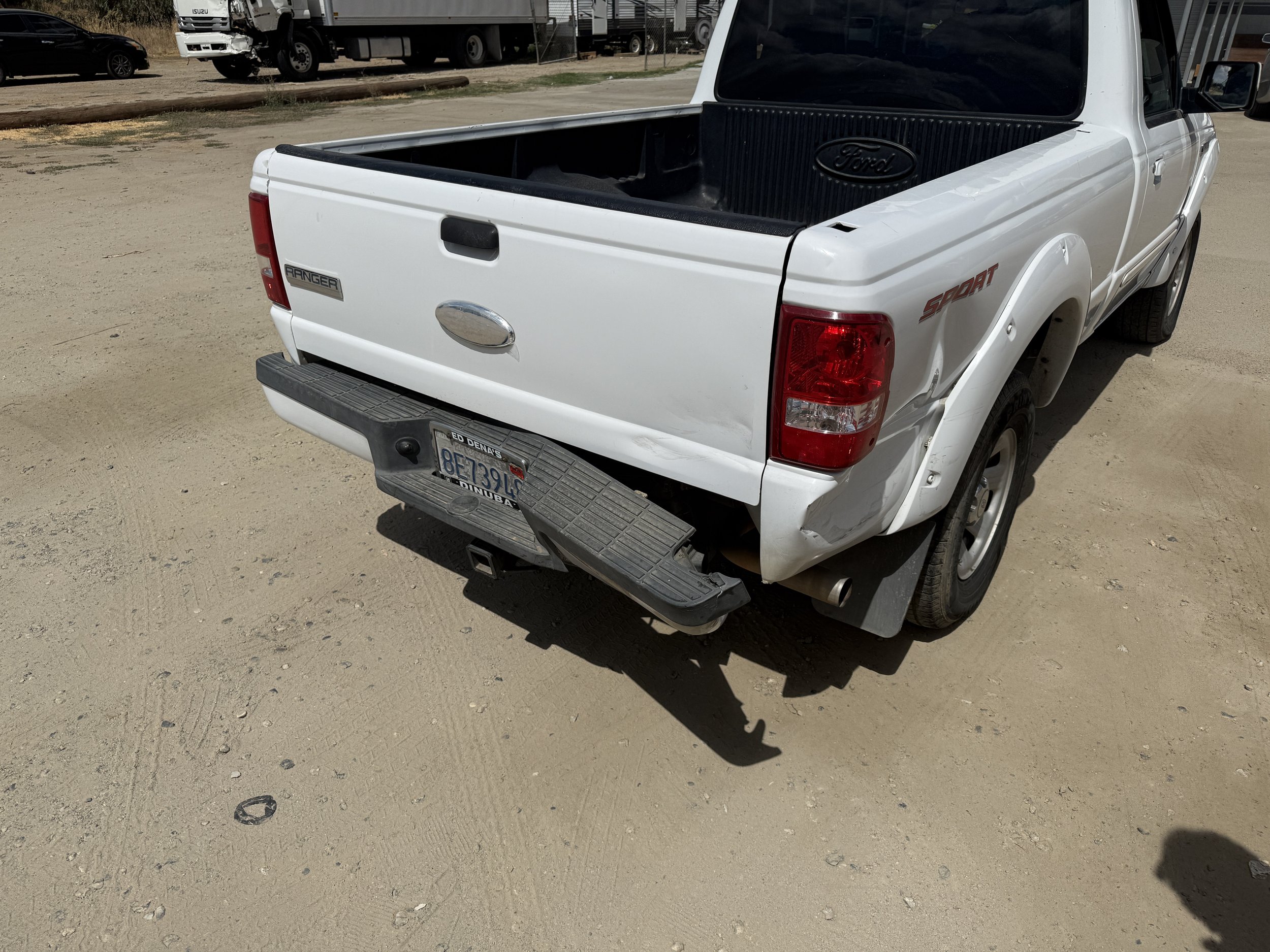 White Ford Ranger pickup truck with a black bed liner, parked on a dirt lot, with a damaged white truck and other vehicles in the background.