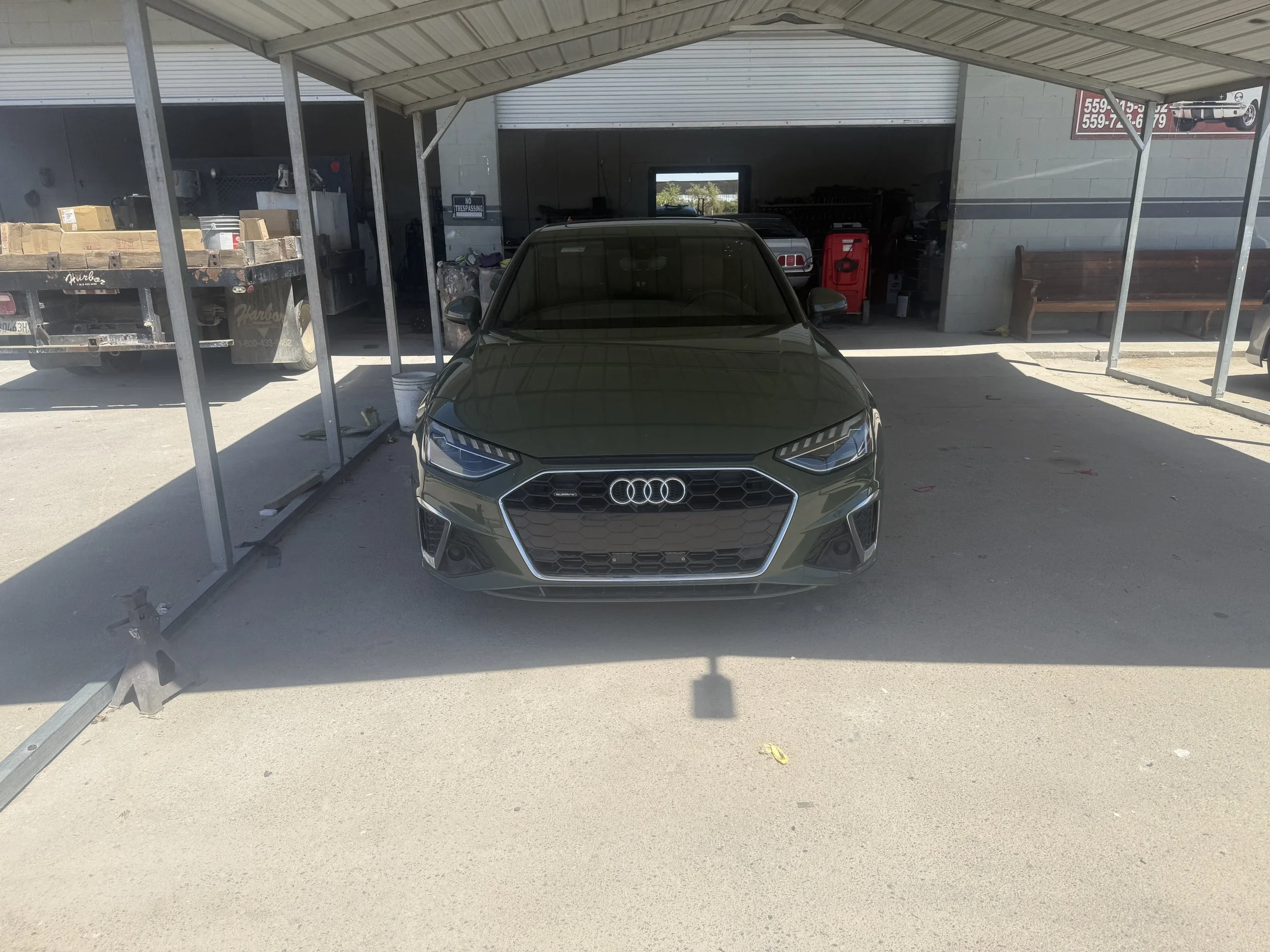 Front view of a dark-colored Audi car parked under a metal carport with a gray wall and garage behind it; there is a wooden bench on the right side and a trailer with boxes on the left side.