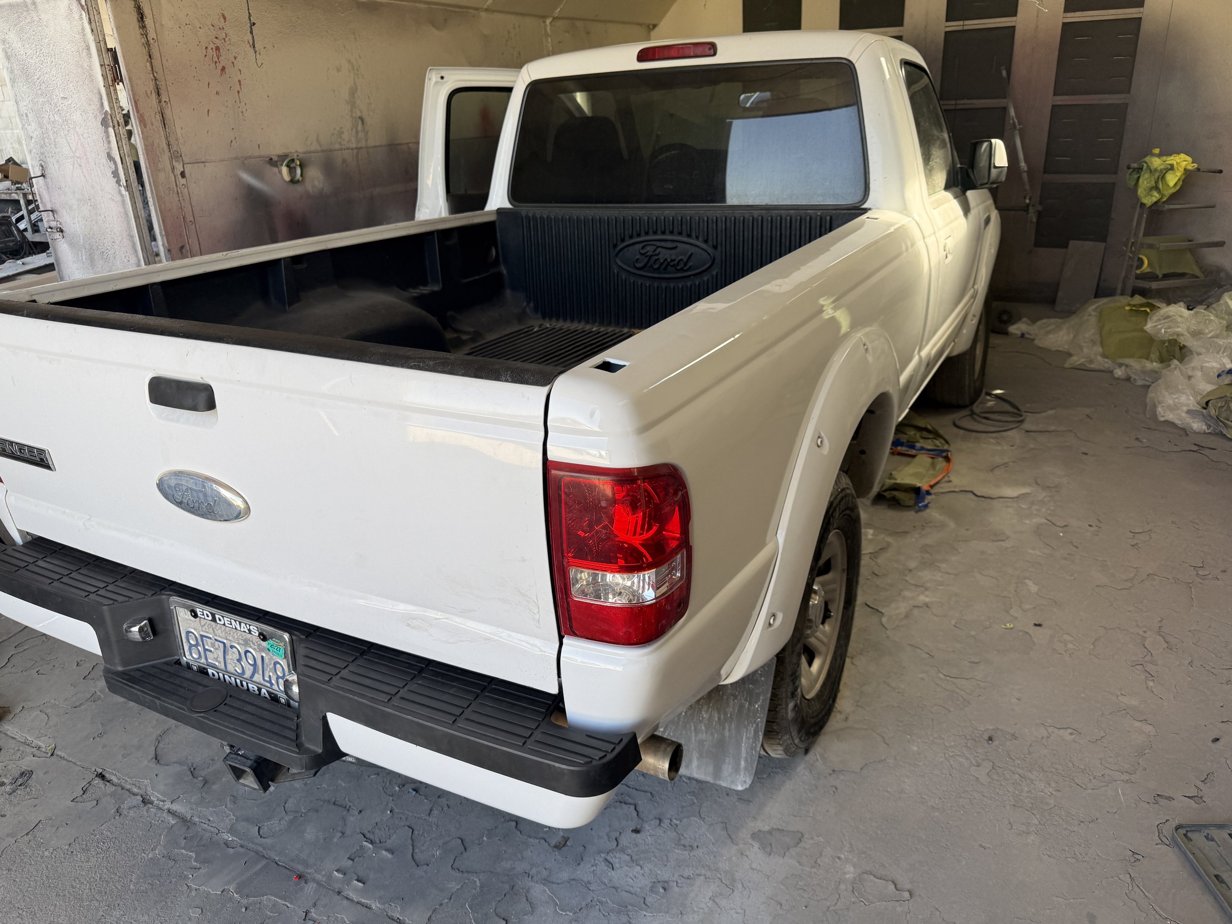 White Ford pickup truck parked inside a garage or workshop, with the tailgate closed and building tools and items visible in the background.