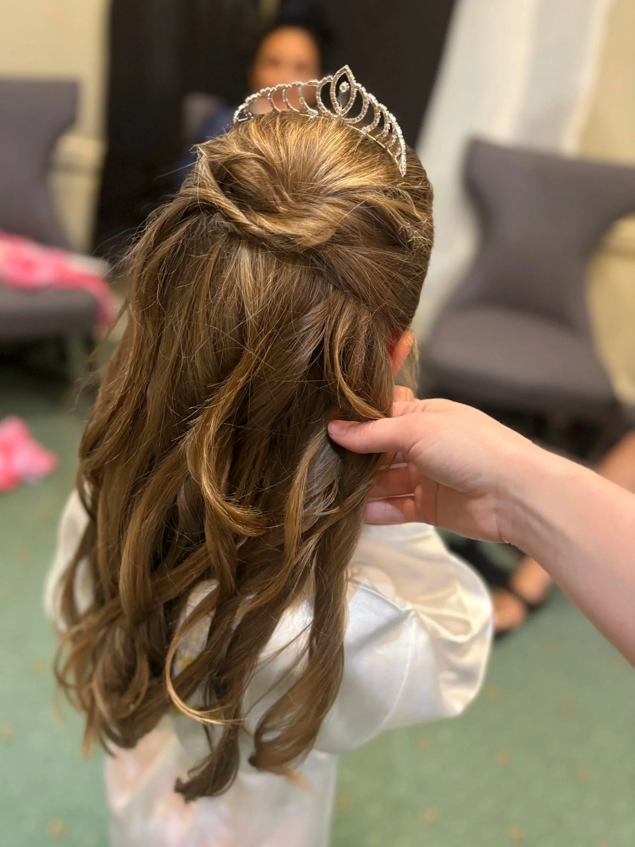 A young girl wearing a tiara and a white dress, with her hair styled in curls, is being attended to by an adult.