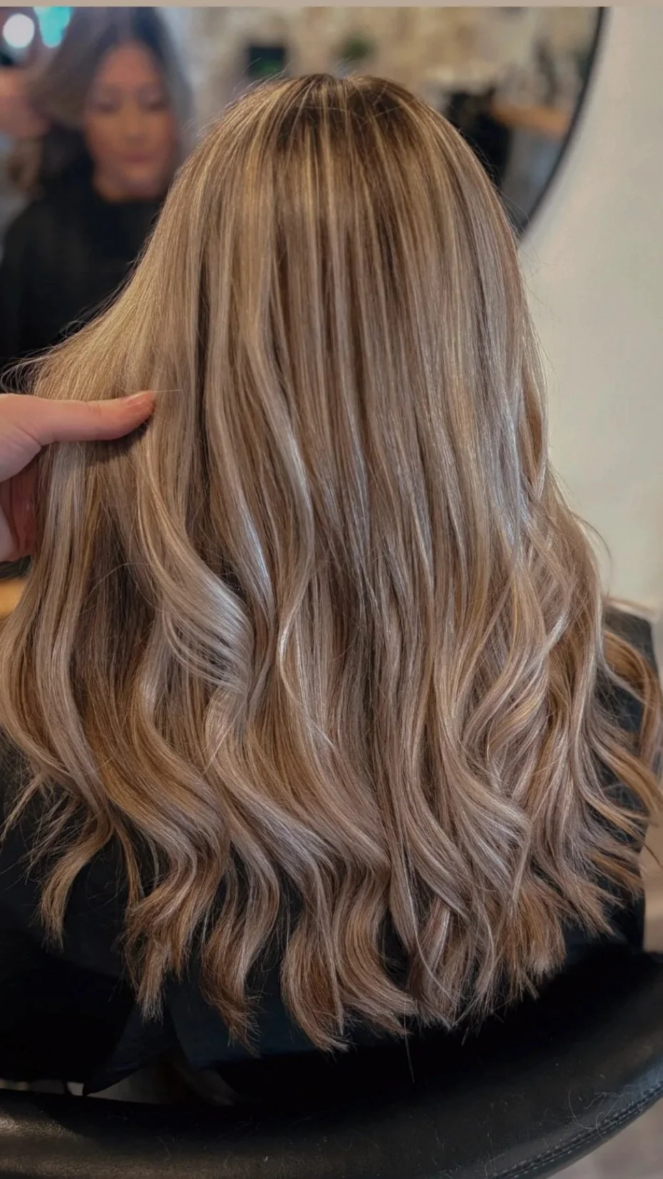 The image shows the back of a woman's head with long, wavy blonde hair. A hand is touching the hair on the left side, and a mirror in the background reflects a woman's face with dark hair.