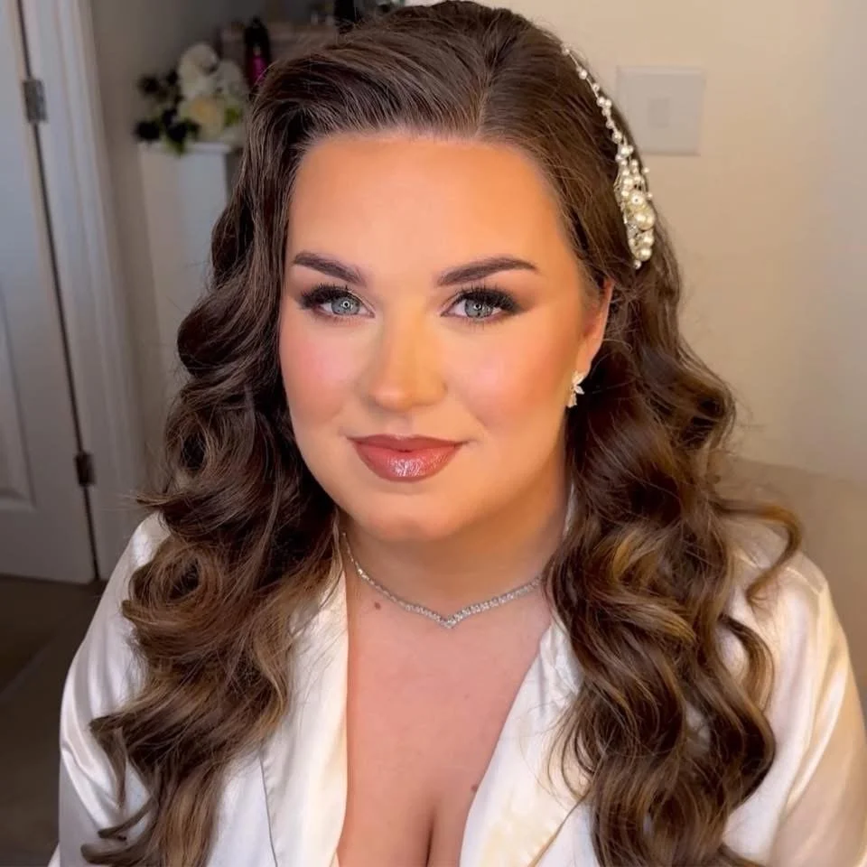 A woman with long, wavy brown hair wearing pearl earrings, a pearl headband, and jewelry, dressed in a satin robe, smiling at the camera in a softly lit room.