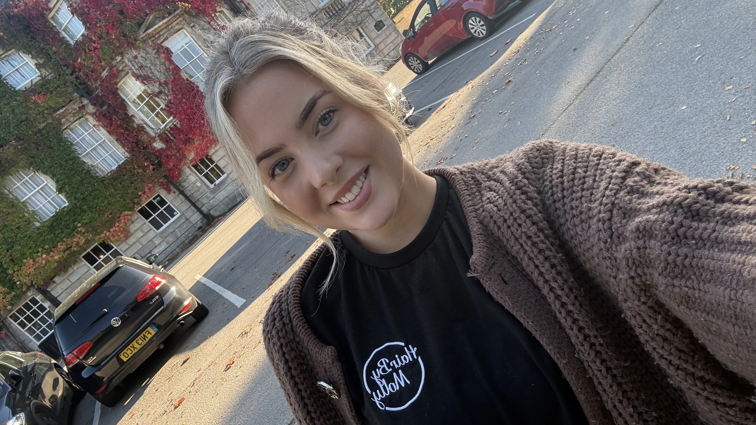 A young woman with blonde hair, blue eyes, and smiling face taking a selfie outdoors, with a building covered in red and green foliage in the background, parked cars on the street, and sunset lighting.